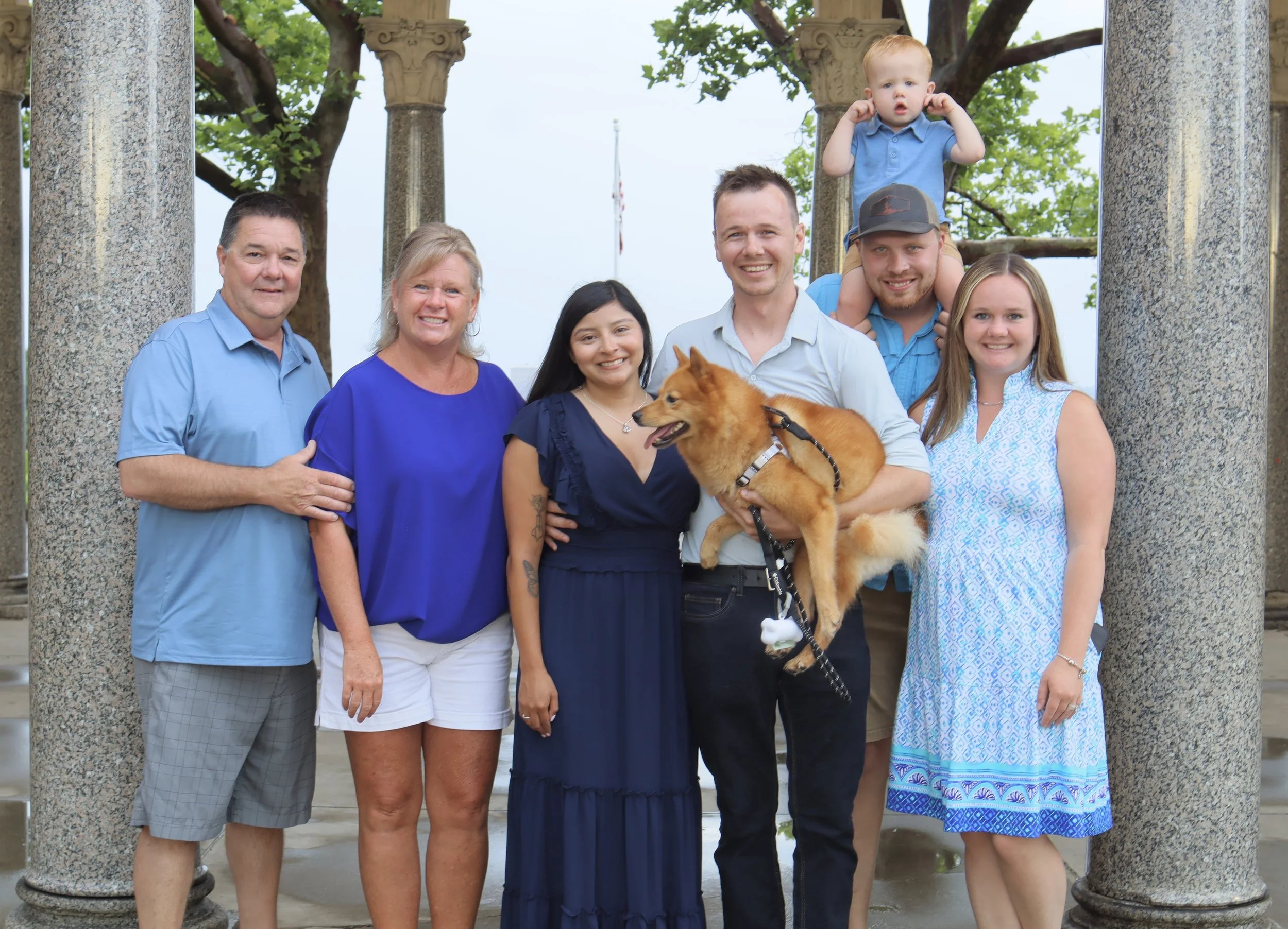 Group photo of eight people and a dog standing outdoors by stone columns with trees in the background. The group includes three men, three women, two boys, and one girl, all smiling and dressed casually.