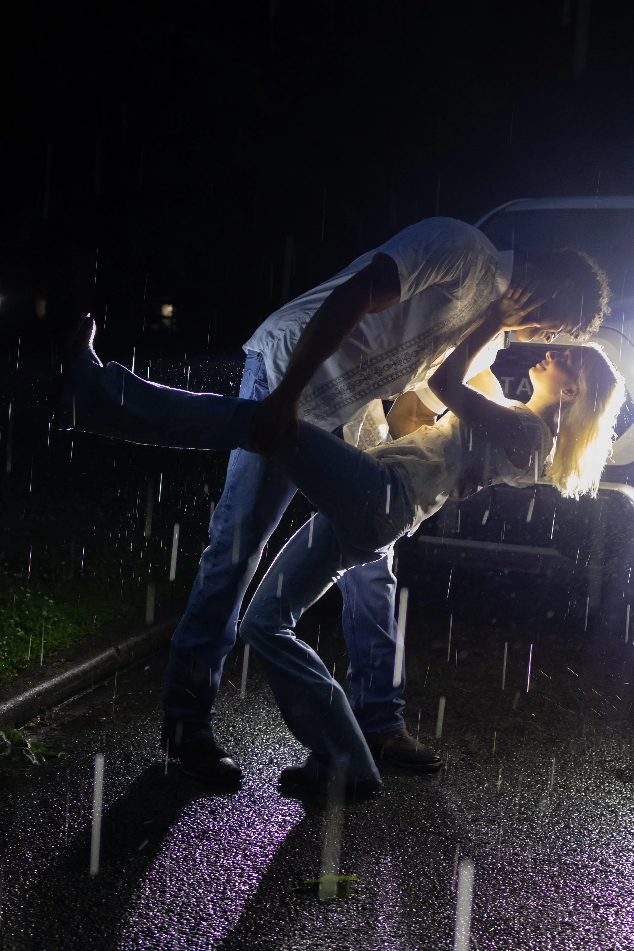 A couple dancing closely outdoors at night during a rainstorm, illuminated by a bright light behind them, with raindrops visible and a wet, reflective street surface.