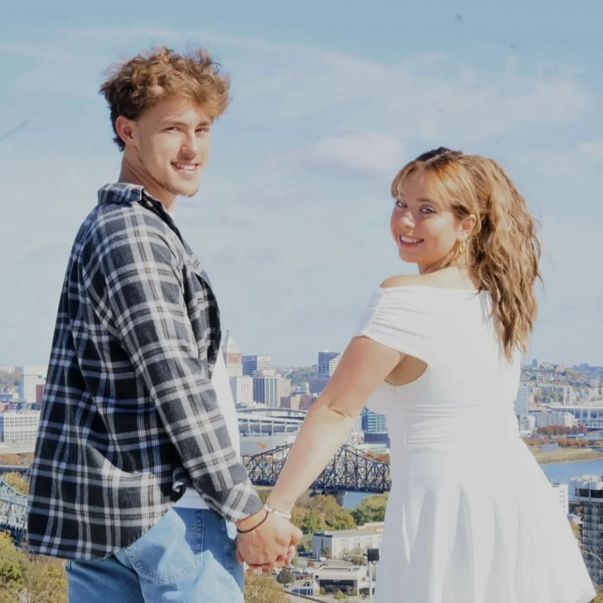 A young couple holding hands and looking at the camera against a city skyline, with a bridge and river visible in the background.