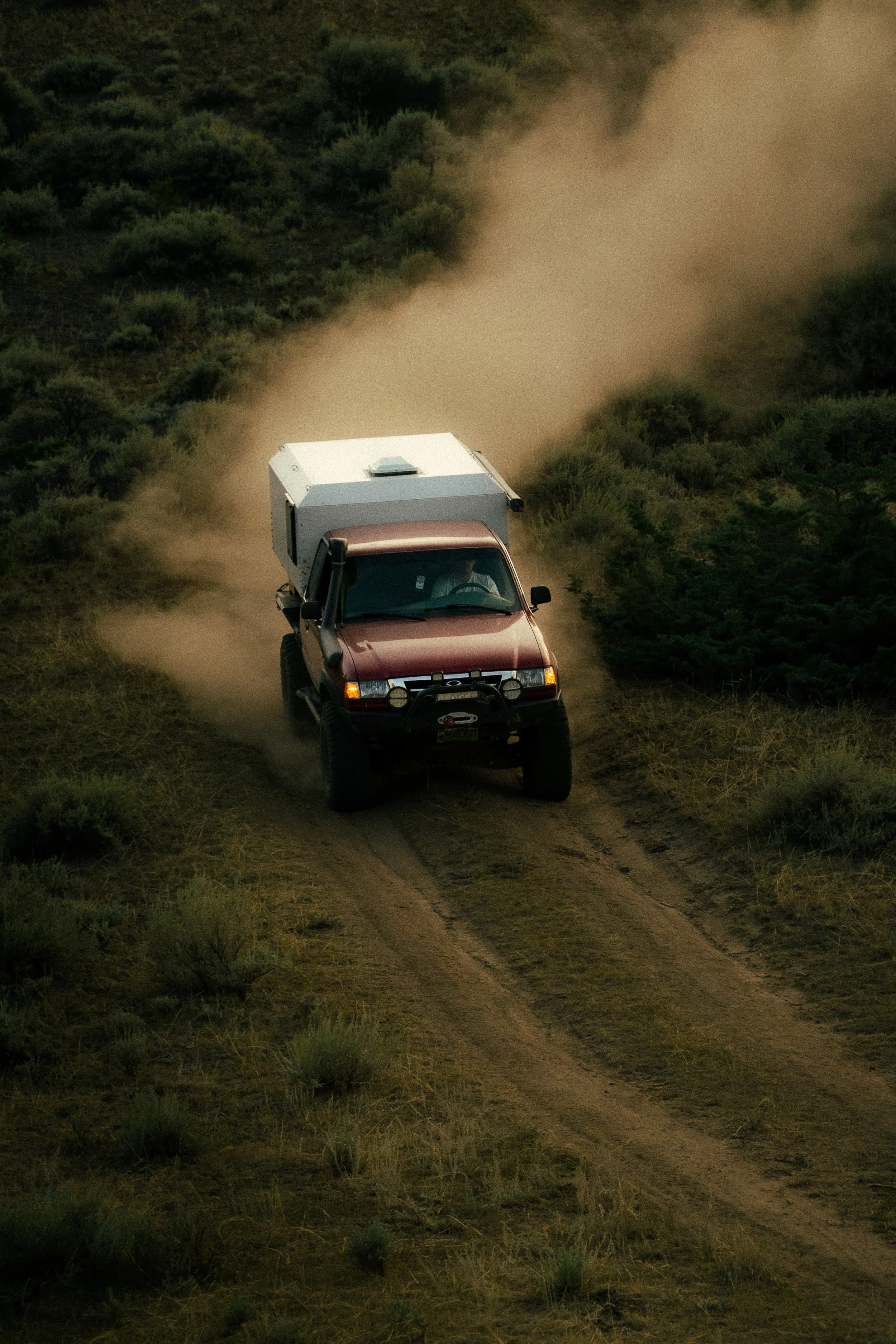 A red off-road vehicle kicking up dust as it drives down a dirt trail through a grassy and shrub-covered landscape.