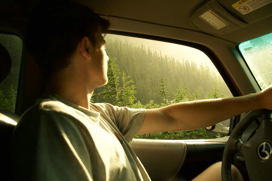 Young man driving a pickup truck through a forested area with tall trees and sunlight streaming in.