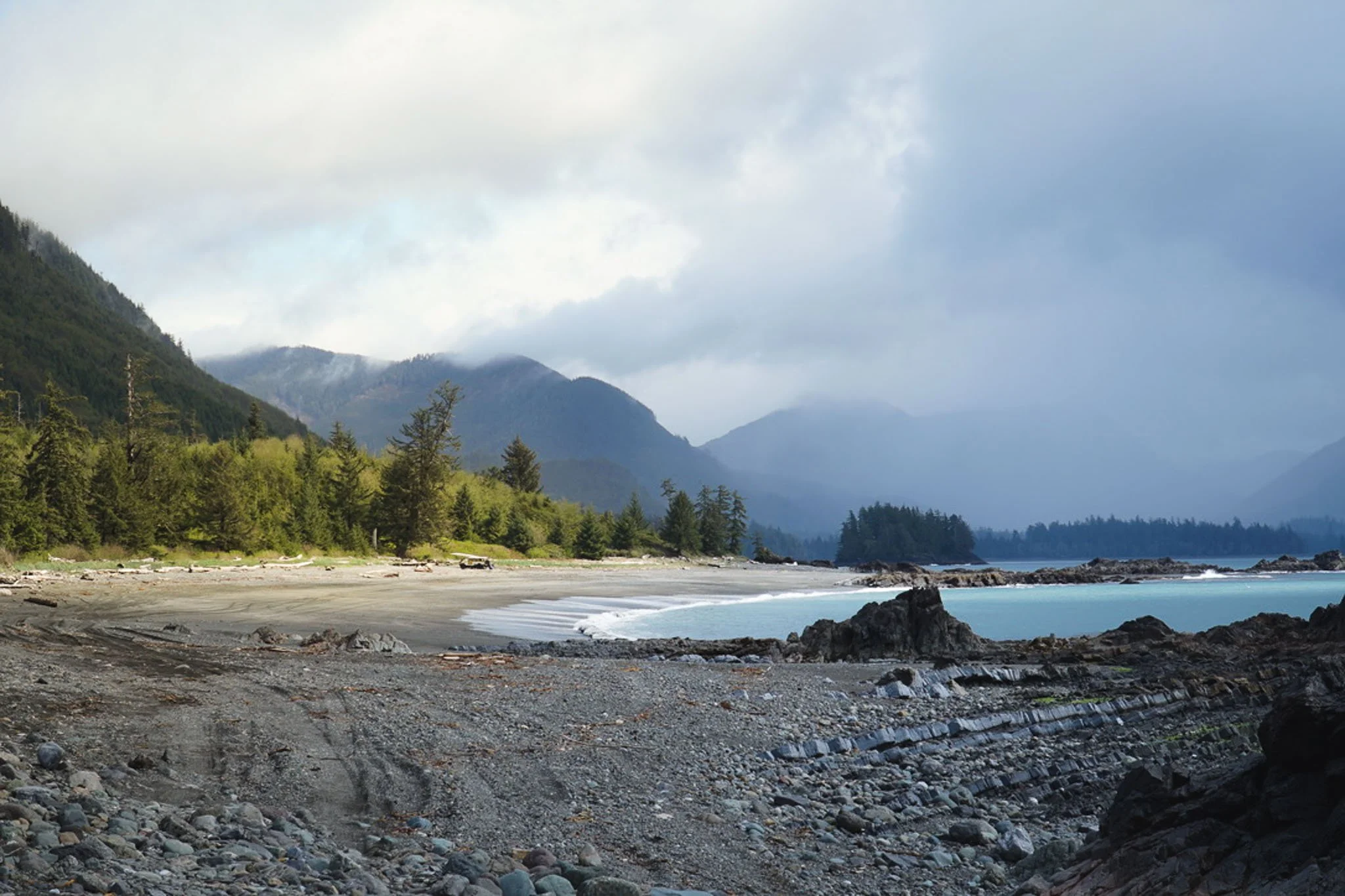 A scenic view of a beach with rocks and driftwood in the foreground, lush green forested mountains in the background, and a cloudy sky over the ocean.