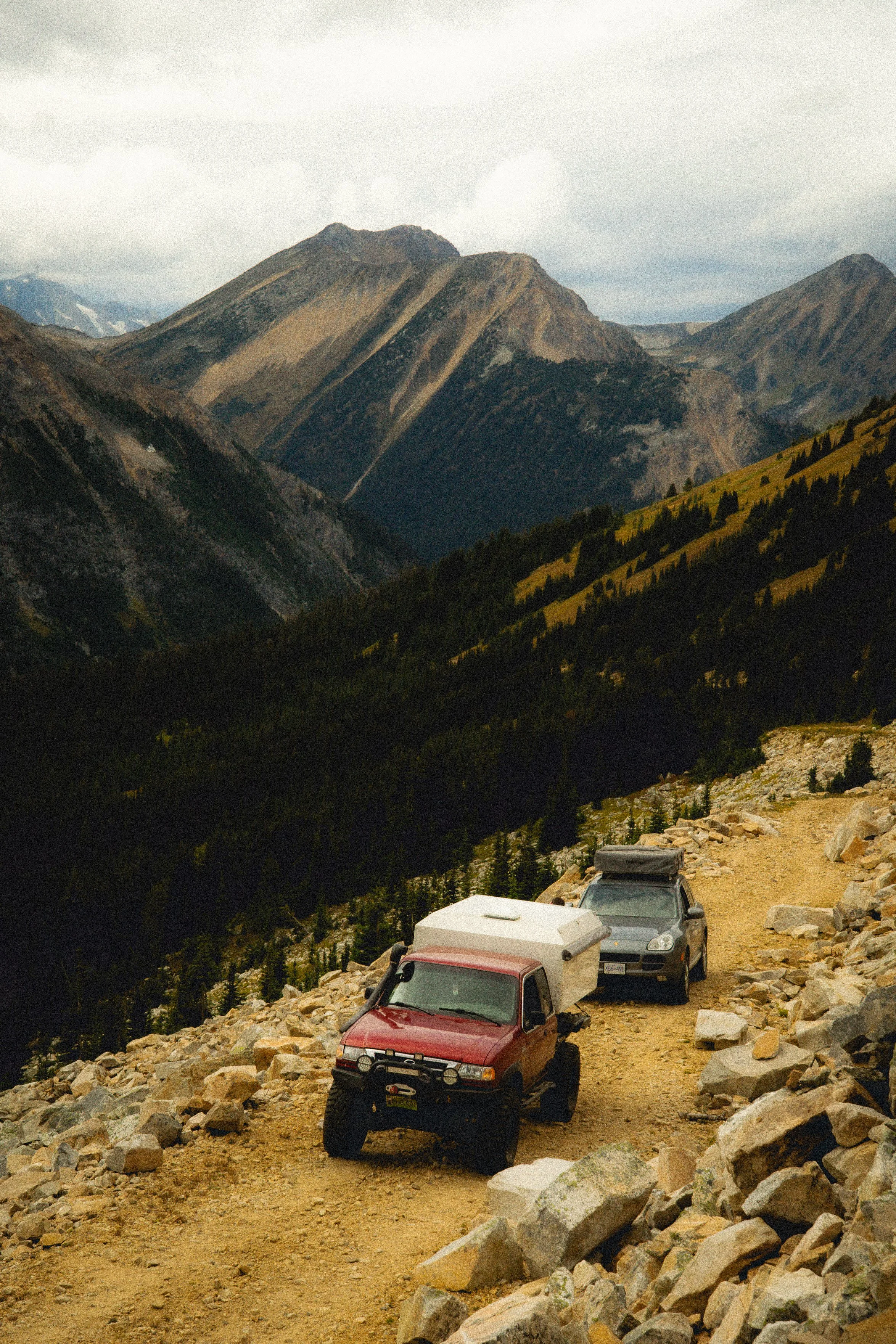 Two off-road vehicles parked on a rugged mountain trail surrounded by large rocks and mountains in the background with cloudy skies.