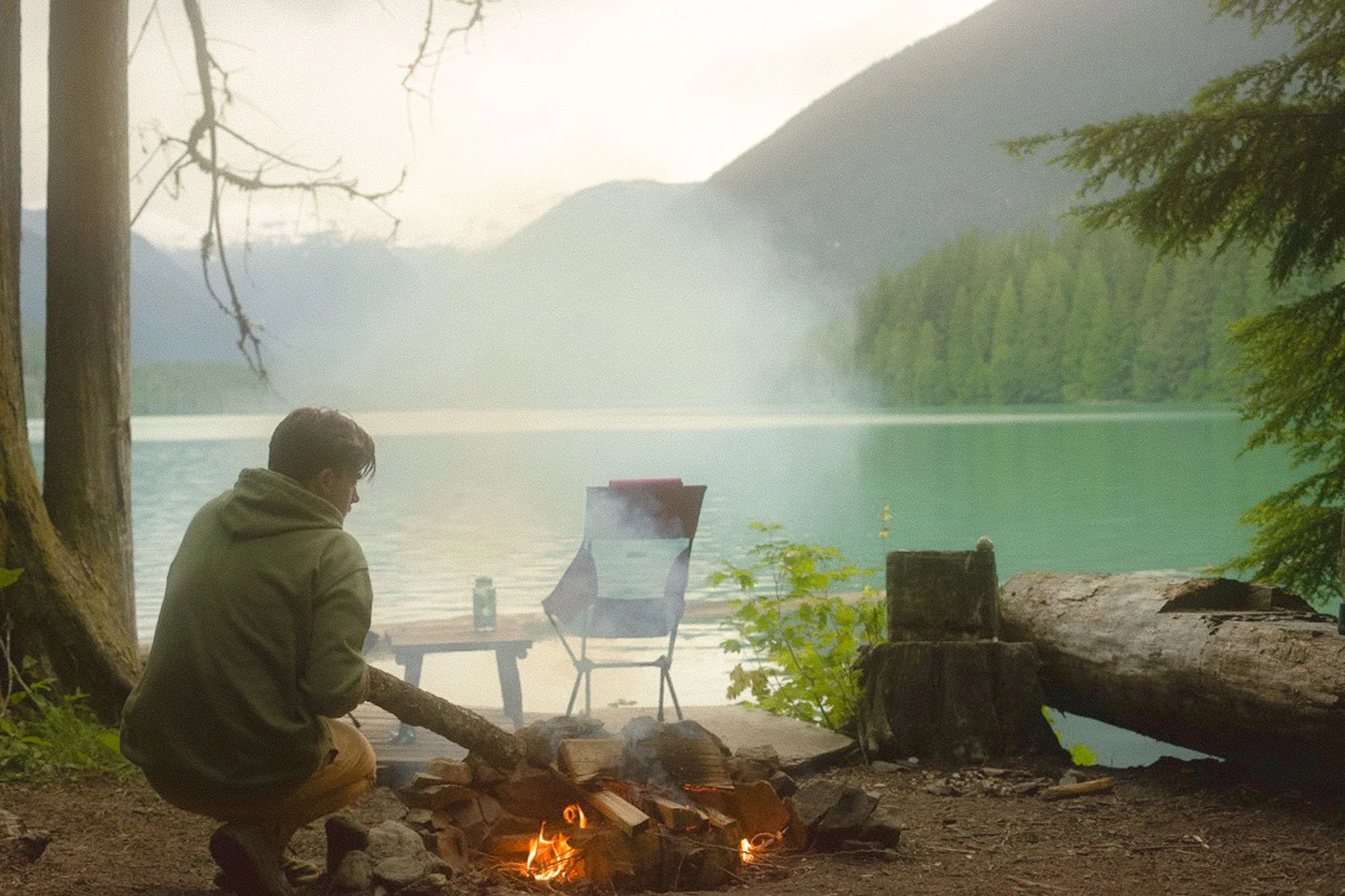 A person sitting by a campfire near a lake, with mountains and trees in the background, on an overcast day.