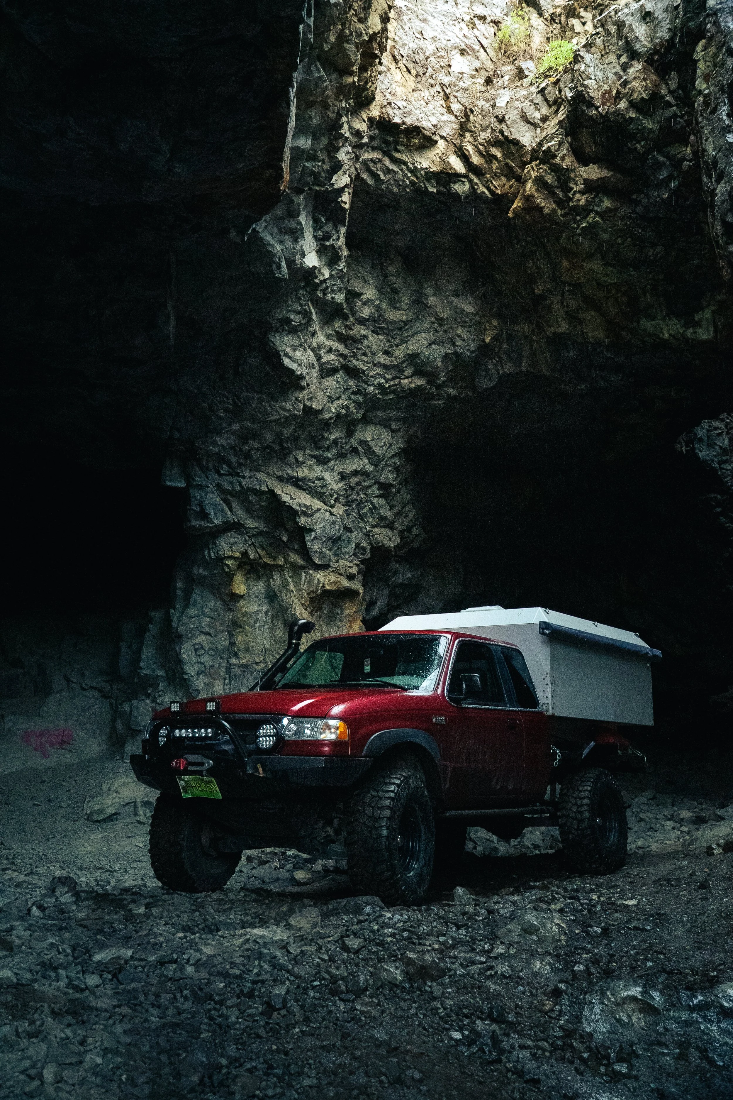 Red off-road truck with a white cargo area parked inside a dark cave with rocky walls and a small opening at the top letting in light.
