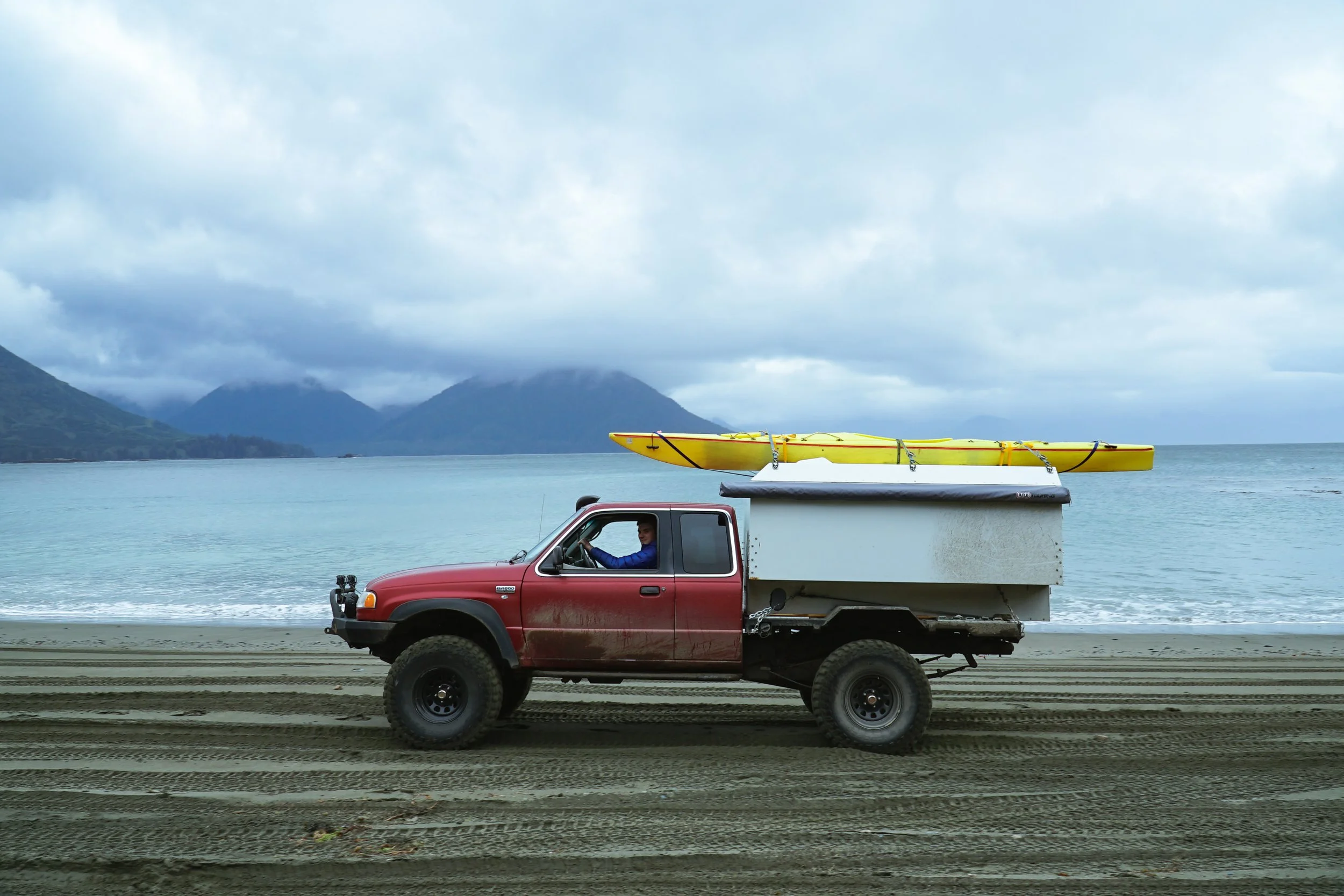 A red pickup truck parked near a beach with a white boat trailer carrying a yellow kayak on a cloudy day, with mountains in the background.