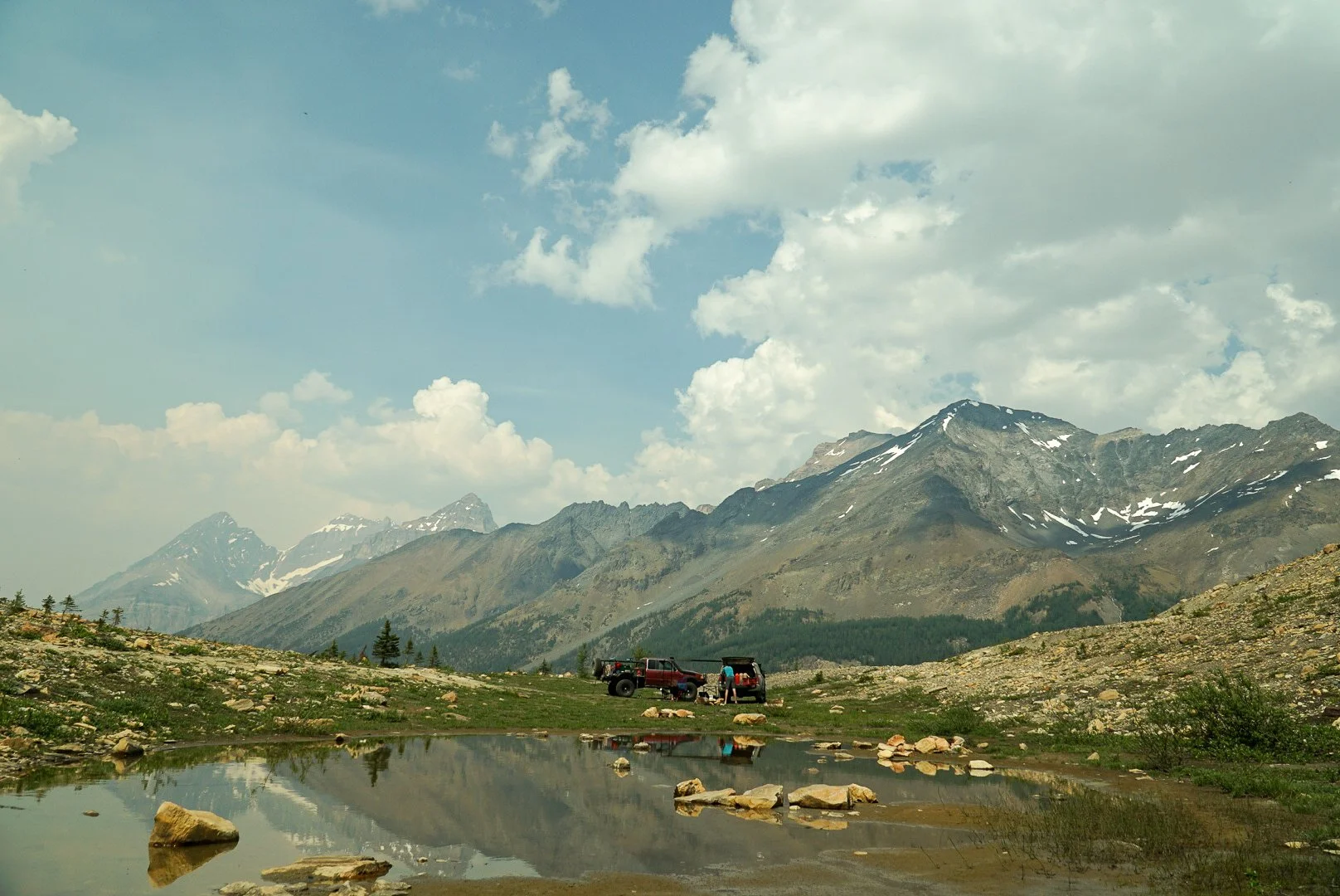 Mountains with snow patches, cloudy sky, two off-road vehicles parked on grassy area near a small pond with rocks, and trees in the background.