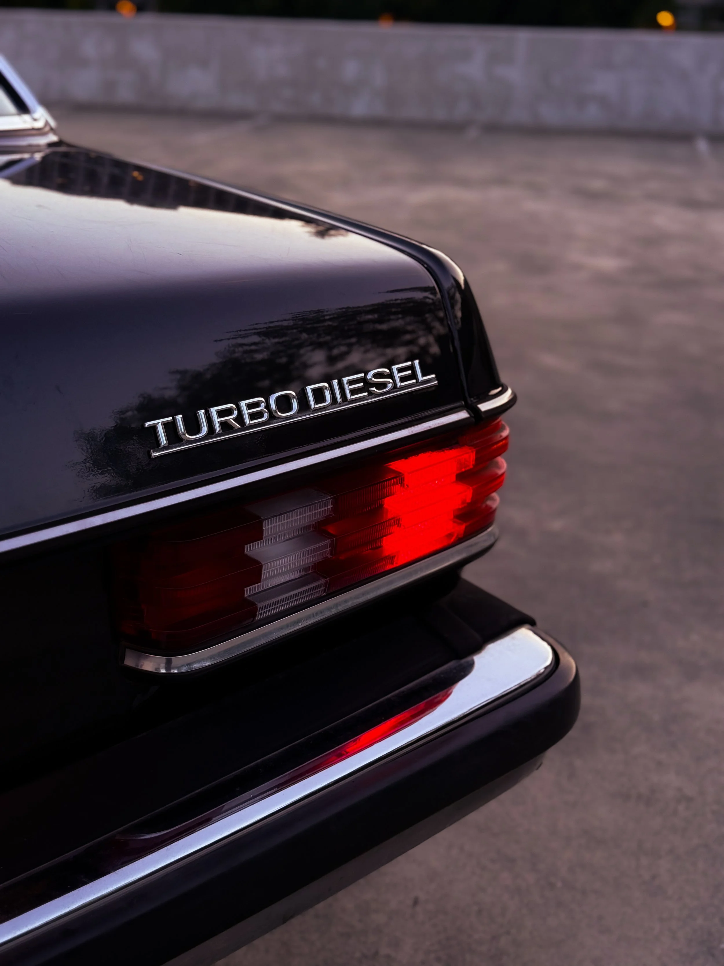 Close-up of a black luxury car's rear with a chrome badge reading 'TURBODIESEL' and a red tail light, parked on a paved surface with a concrete barrier in the background.