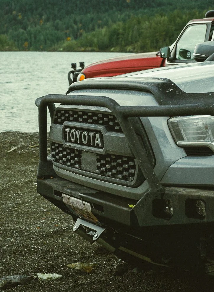 Close-up of the front of a gray Toyota off-road vehicle with a black grille guard, parked on gravel near a body of water with trees in the background.