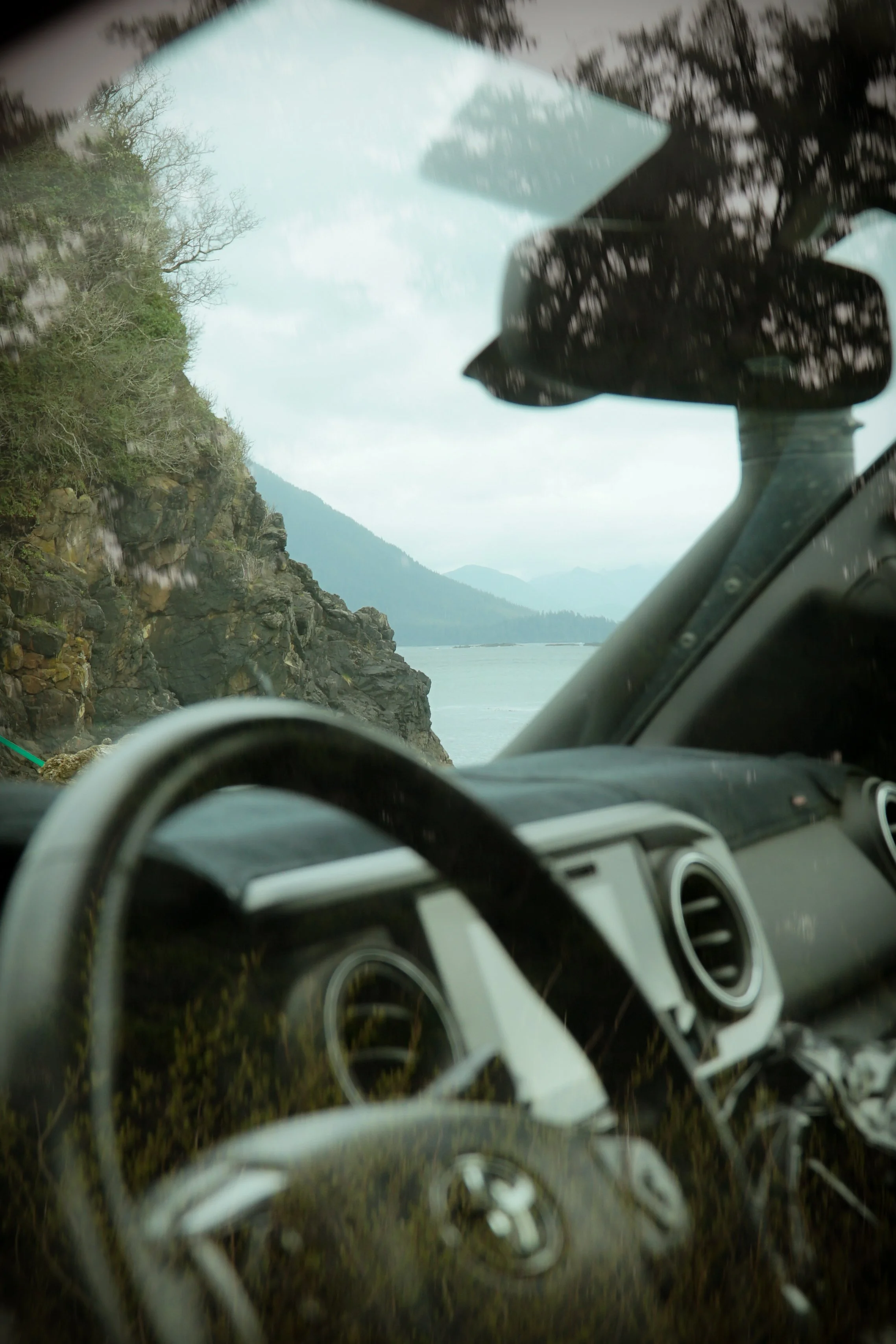 Inside view of a vehicle with reflections of trees and a mountain on the windshield, with a scenic landscape of rocky shoreline, water, and hills in the background.