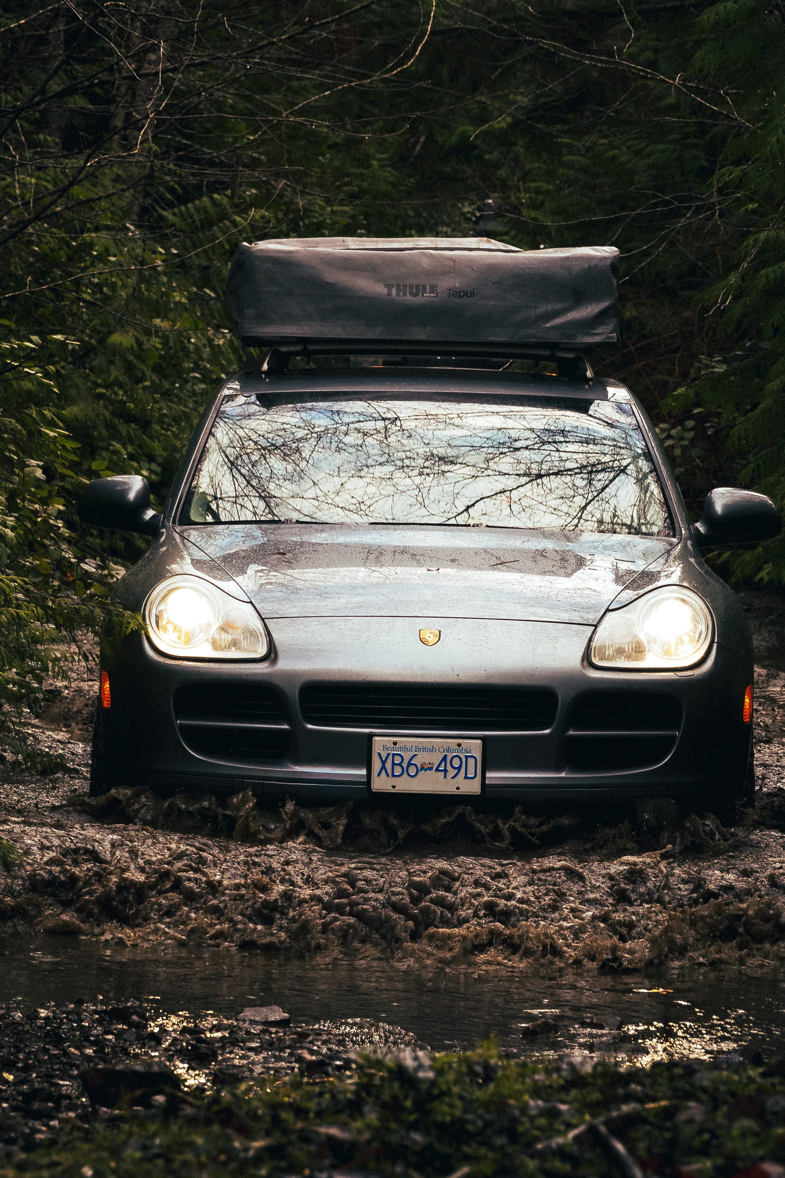A black Porsche car driving through a muddy trail in a forest, with water splashing up around the front tires. The car has a roof cargo box and a British Columbia license plate.
