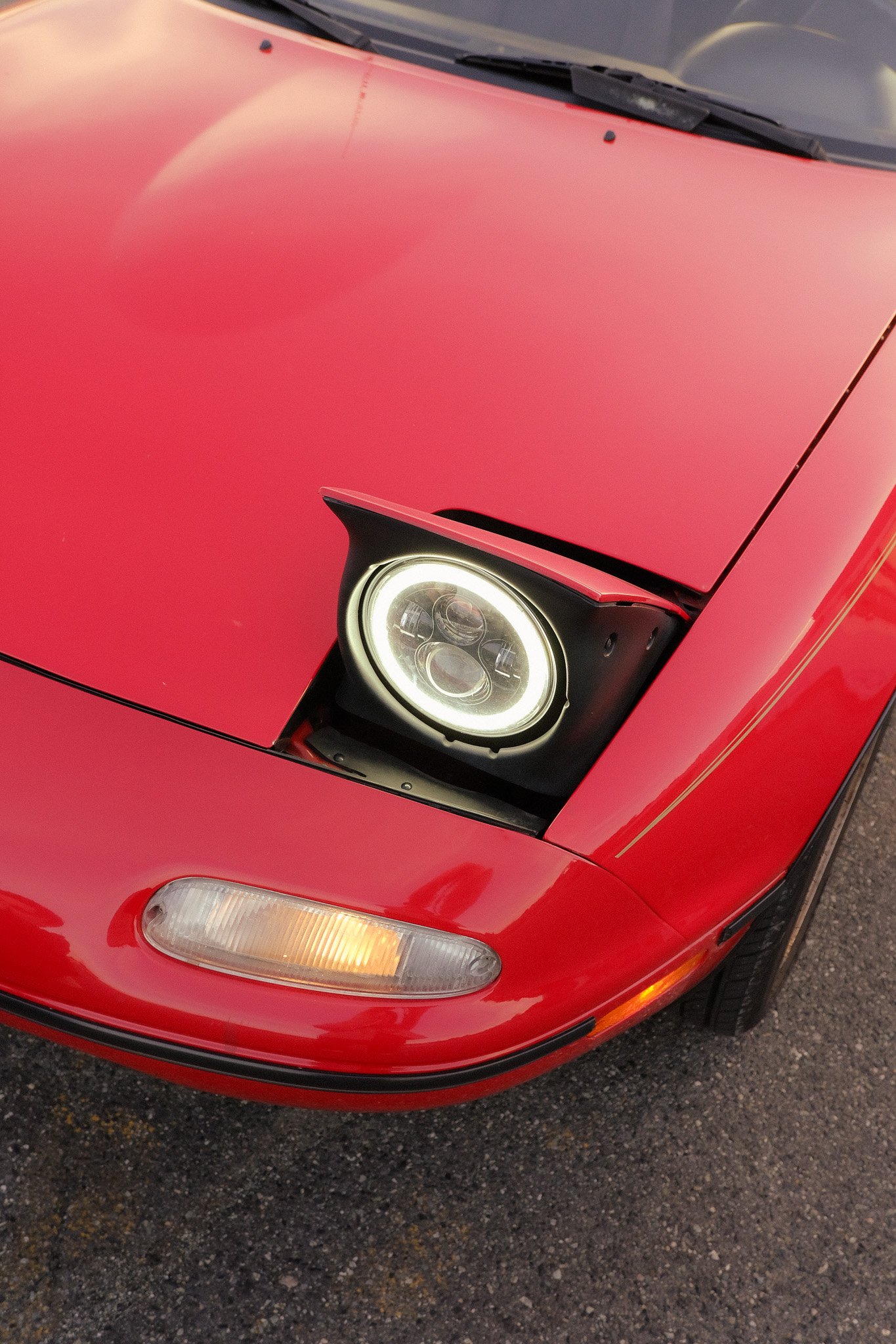 Close-up view of the front right of a red sports car, showing its headlight and part of the hood, parked on a gravel surface.