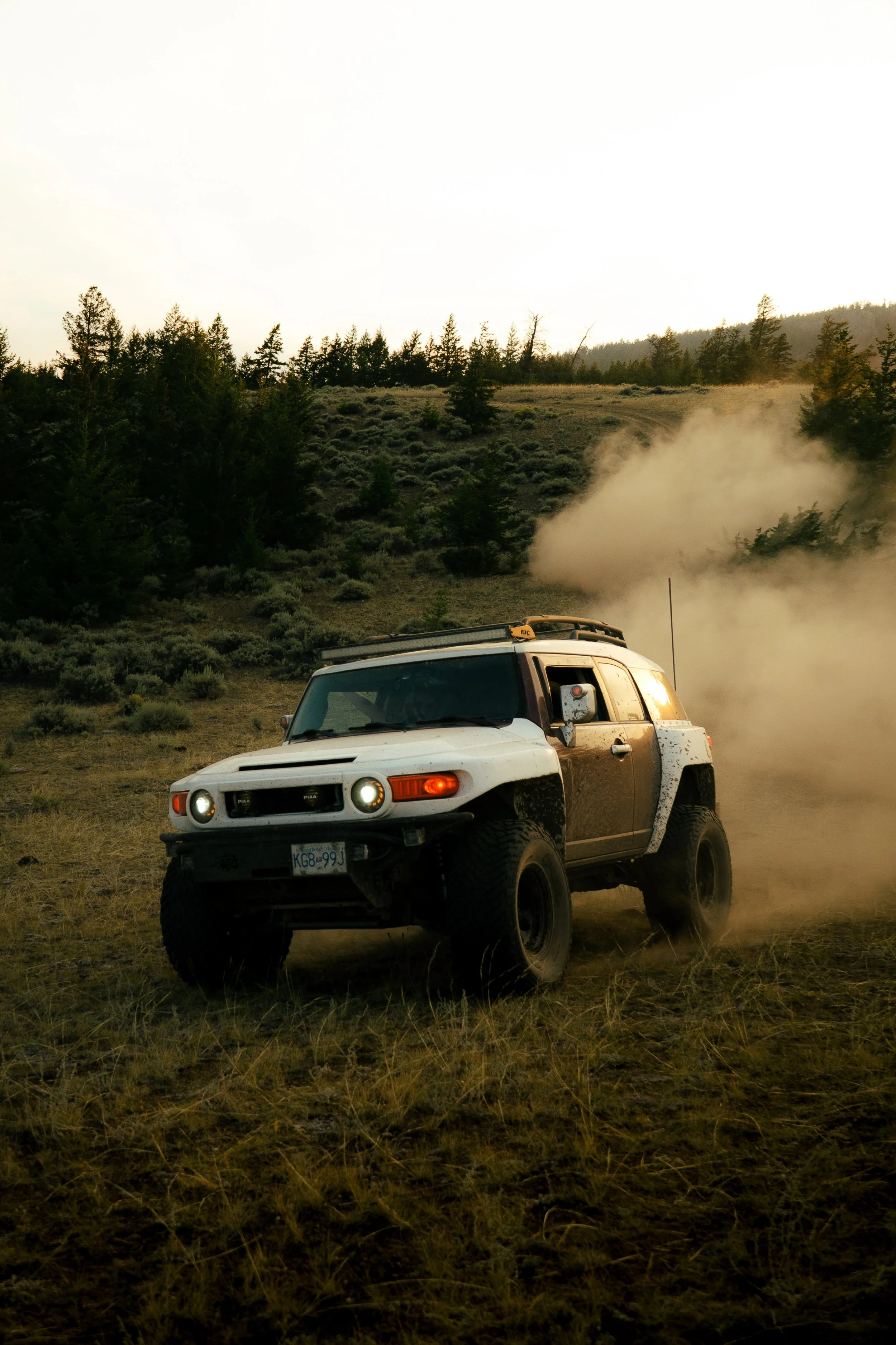 A white off-road vehicle driving through a dusty field with a background of trees and hills, and dust or smoke rising behind it.
