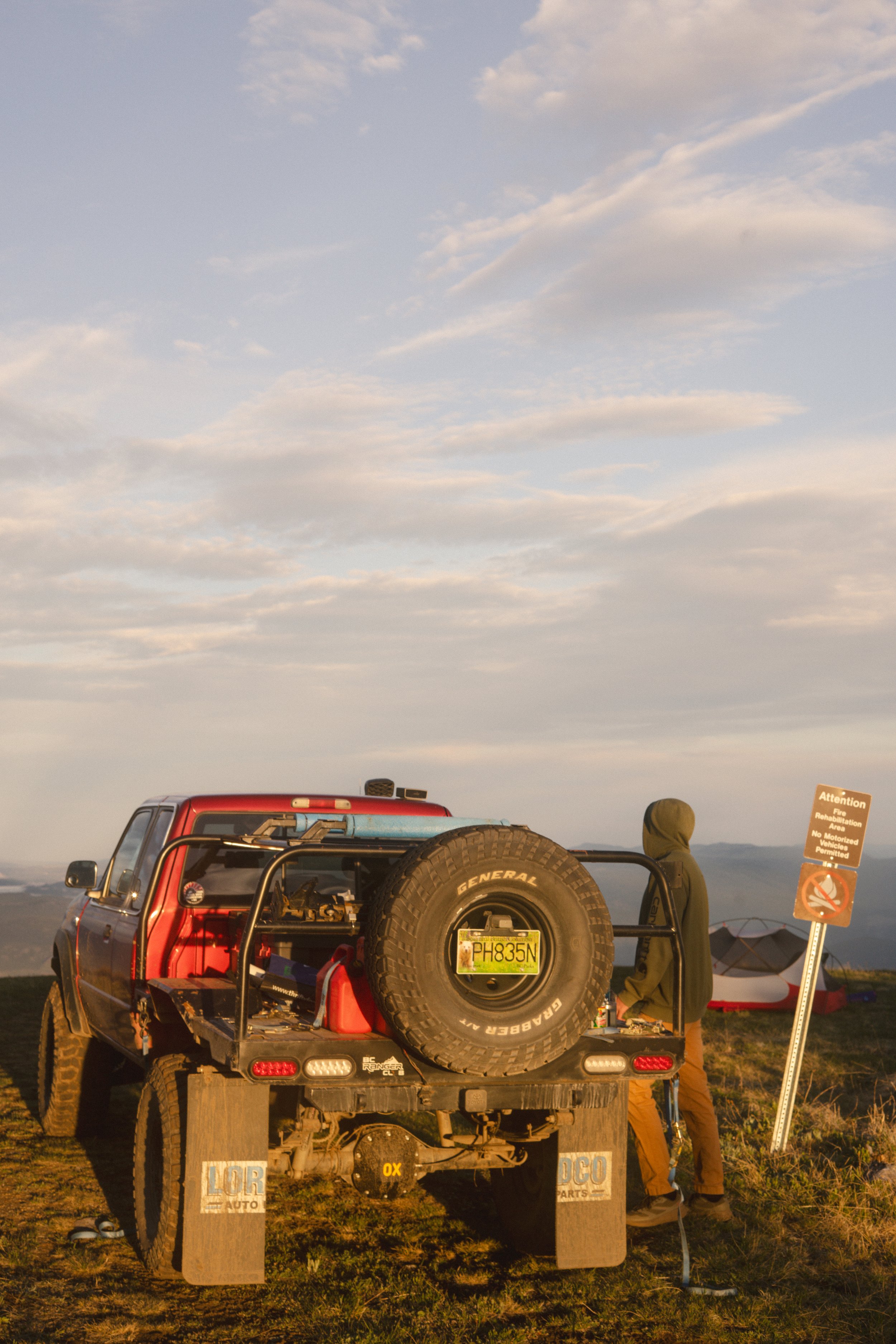 Off-road vehicle with a spare tire mounted on the back, parked on a grassy field at sunset. A person with a hat is standing beside it, and there are signs indicating a designated recreation area.