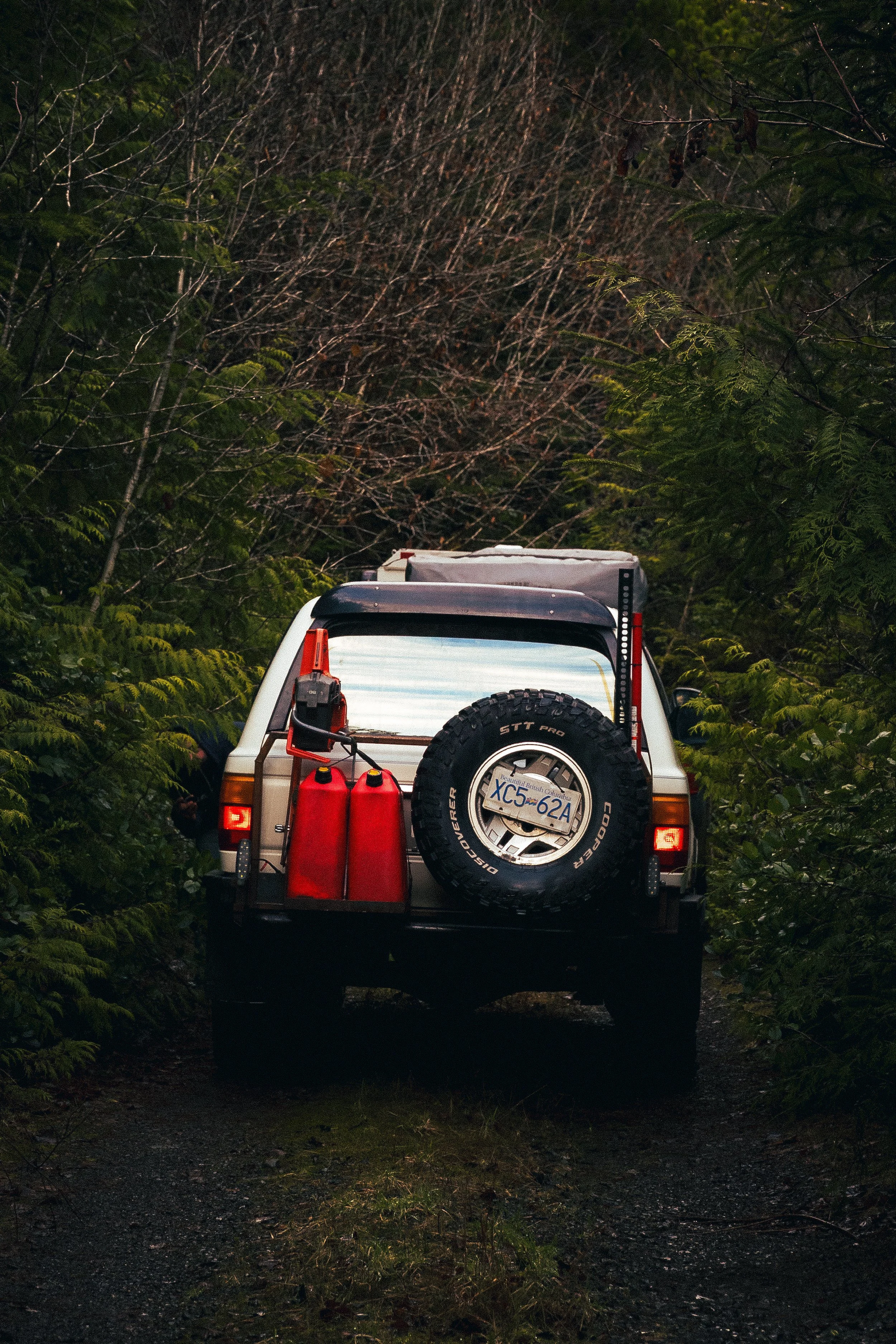 Back view of a white off-road vehicle with a spare tire, two red fuel containers, and a bumper mounted holder, parked on a dirt road surrounded by dense green foliage and trees.