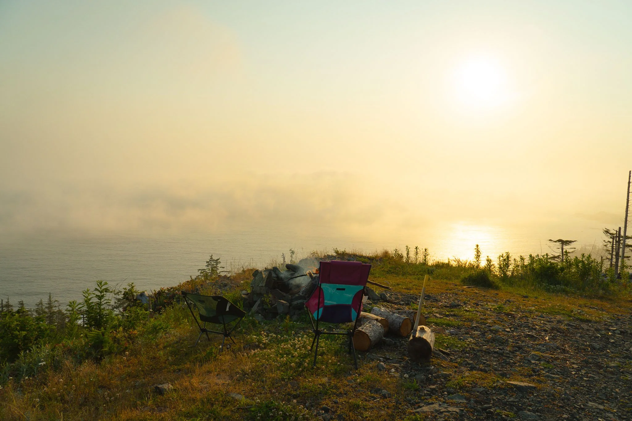 Seaside view at sunset with trees, rocks, and camping chairs on a grassy area facing calm water.