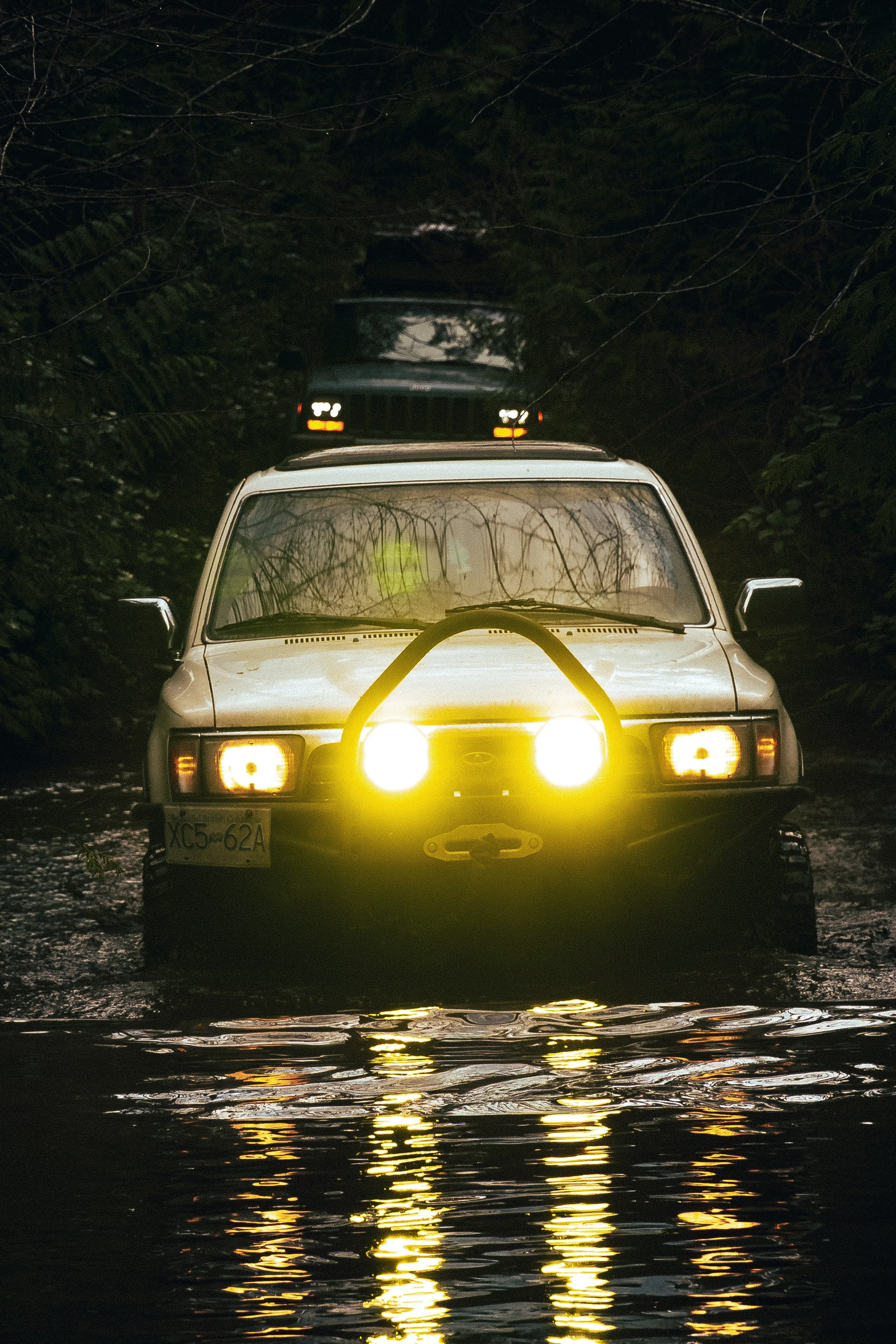 A white vintage car with its headlights on, reflected on water, in a dark outdoor setting surrounded by trees and bushes.