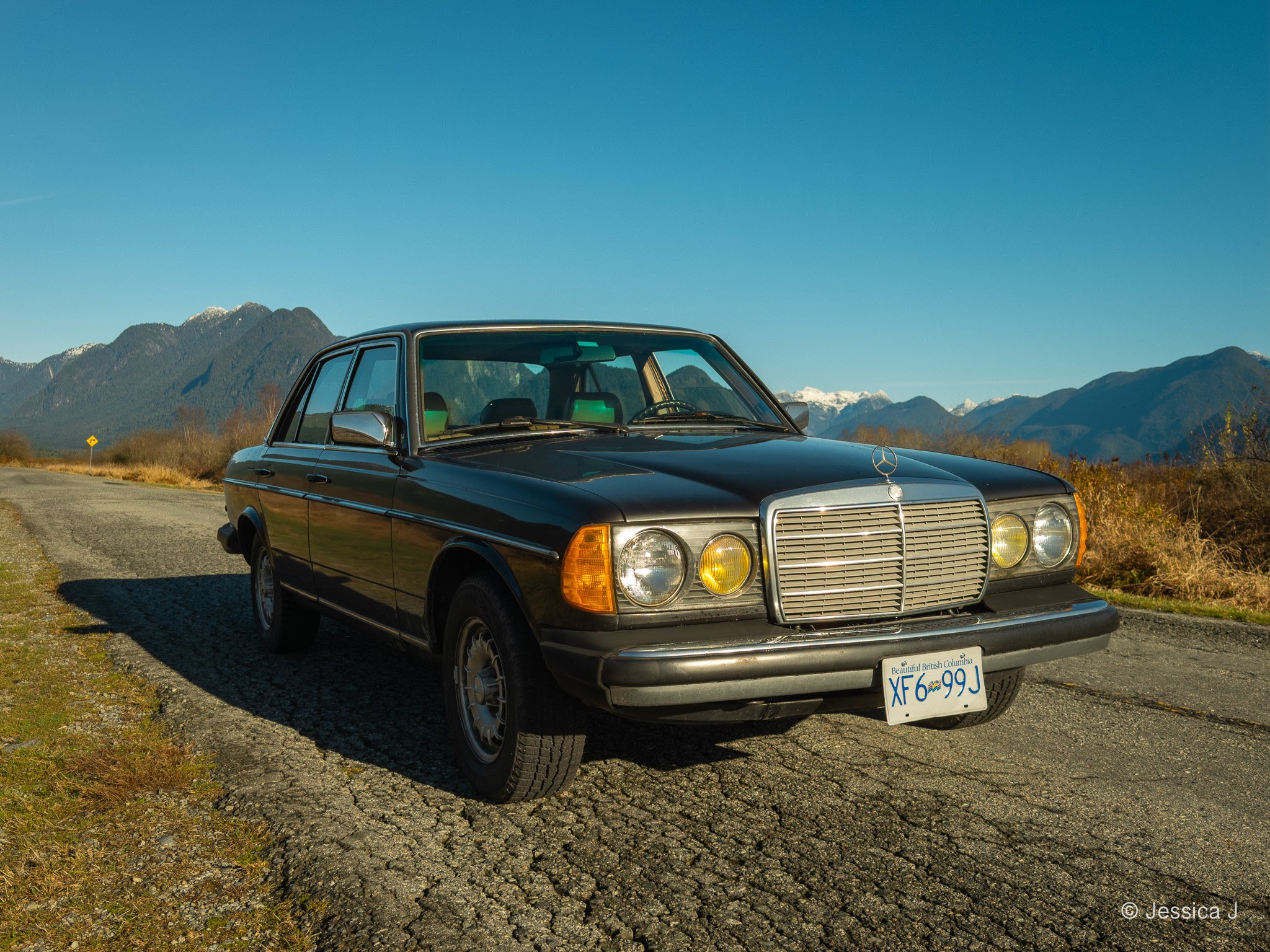 A black vintage Mercedes-Benz car parked on a gravel roadside with a mountainous landscape and clear blue sky in the background.