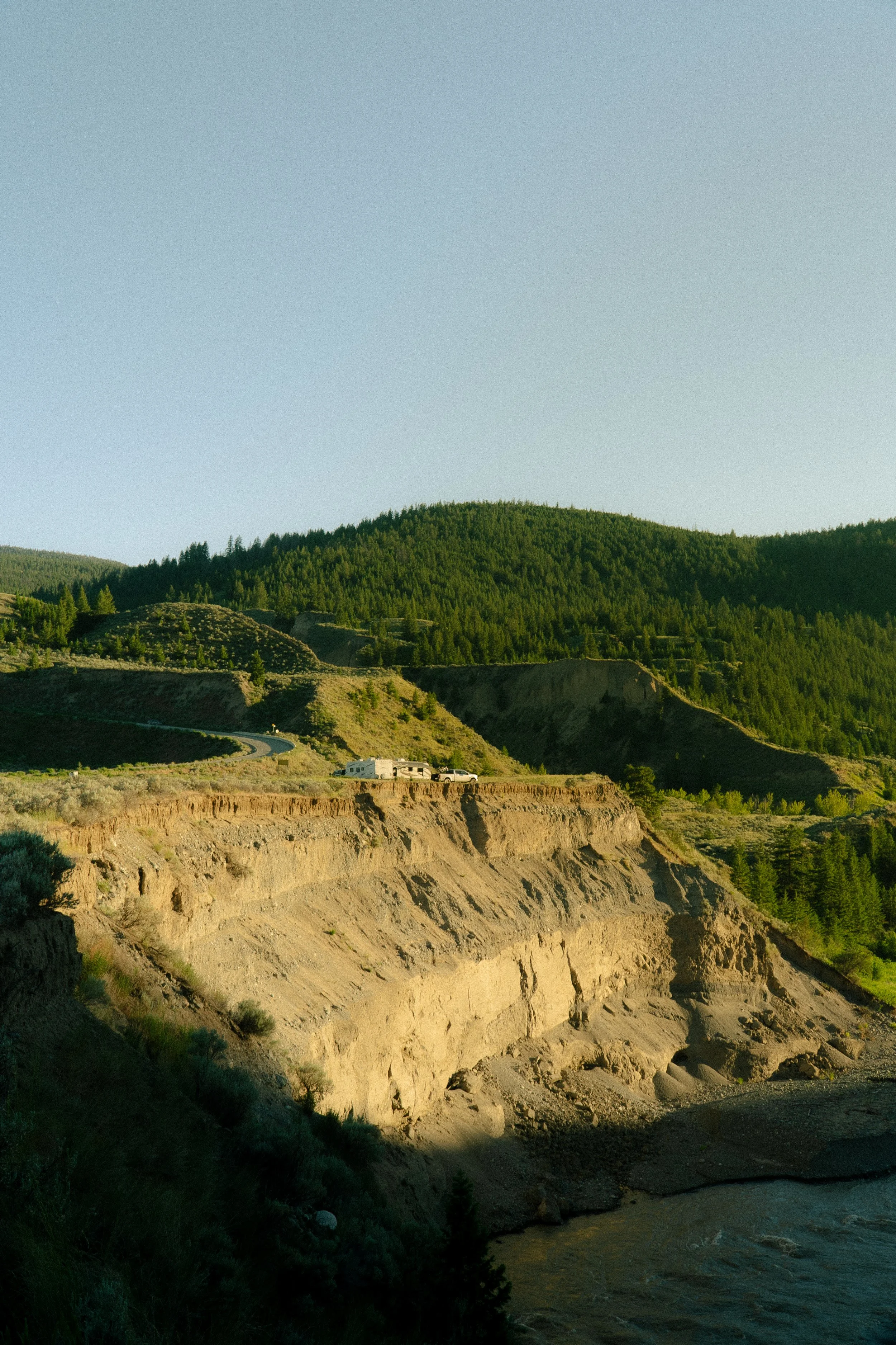 A landscape of green forested hills, a winding road, and a small white building with a vehicle parked near it, set against a clear blue sky.