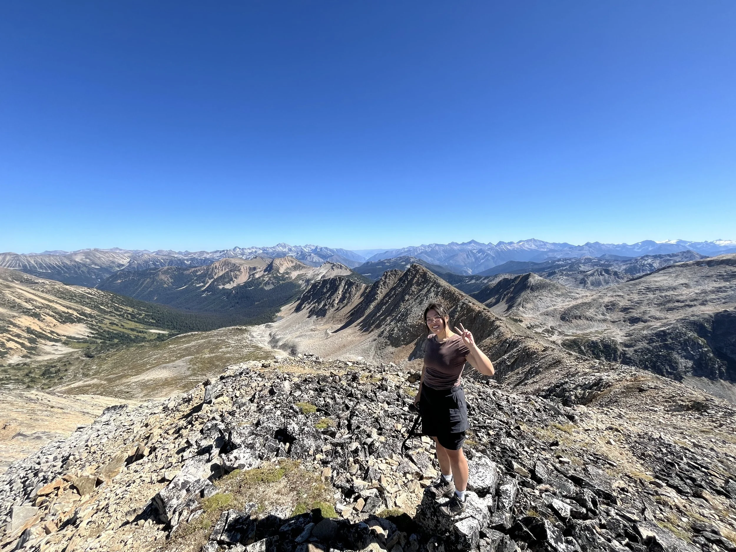 A woman smiling and making a peace sign with her fingers stands on rocky terrain in a mountainous landscape with a clear blue sky, rugged mountains with some snow-capped peaks, and sparse vegetation.