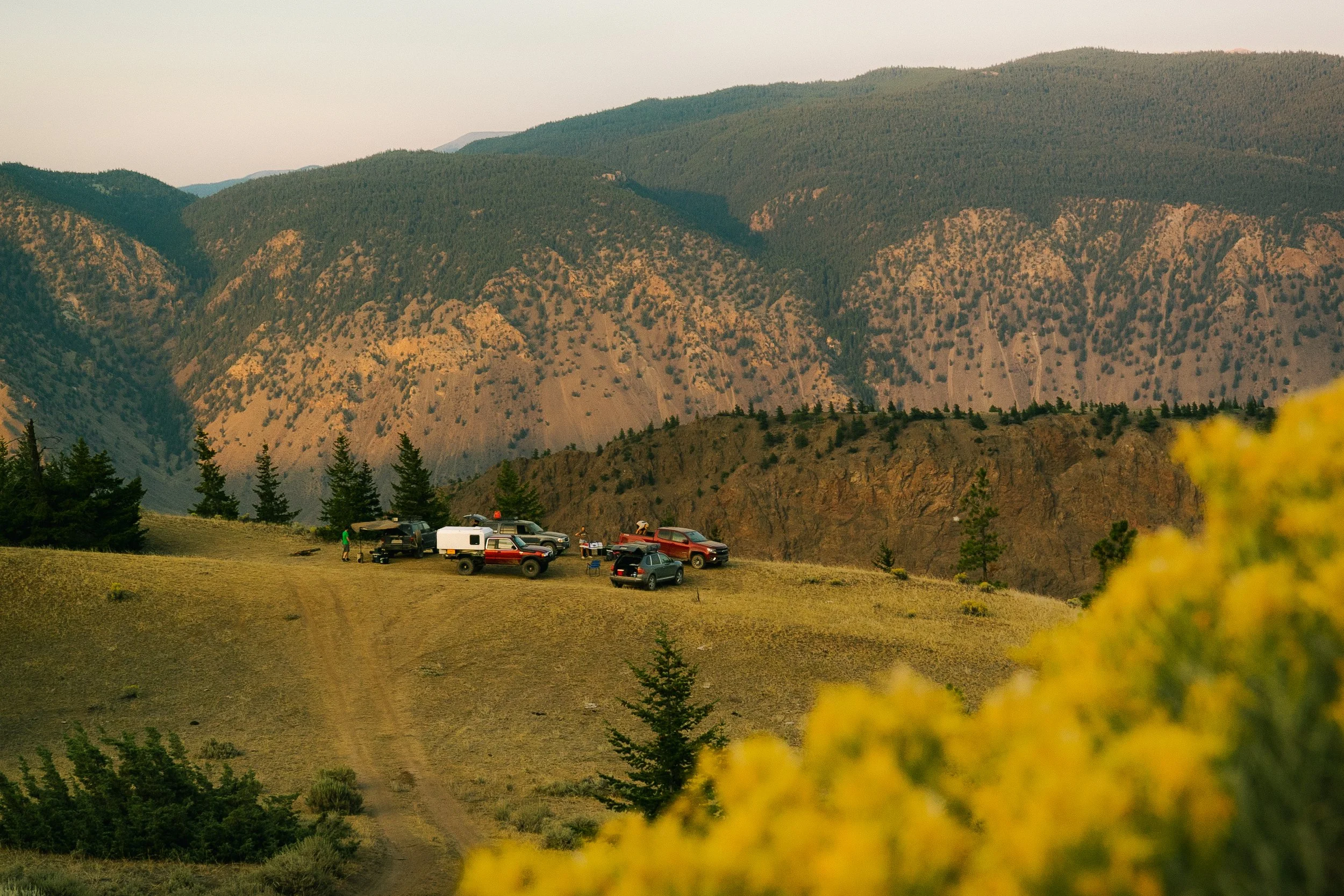 A mountain range with varying vegetation, a small parking area with several cars, and a few people near the vehicles, set against a scenic landscape with distant hills.