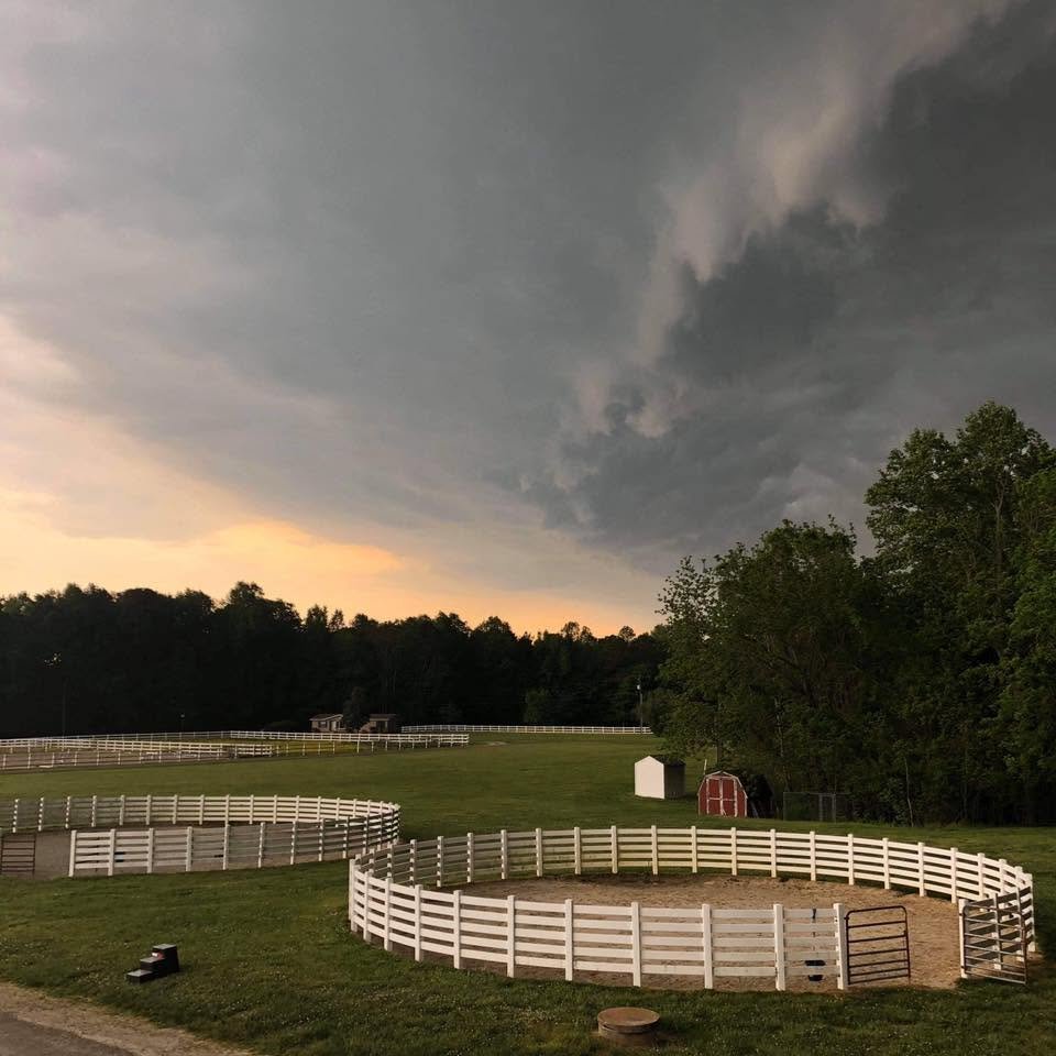 Horse riding arena with white fencing under dark stormy sky, trees and small sheds in background.