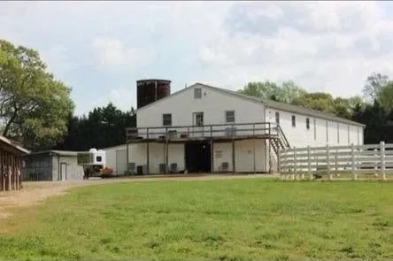 A white barn with a raised walkway and railing, a black chimney, and a grassy field in front.