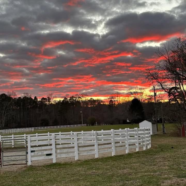 Sunset with vivid red and orange clouds over a farm with white fences, trees, and a small shed.