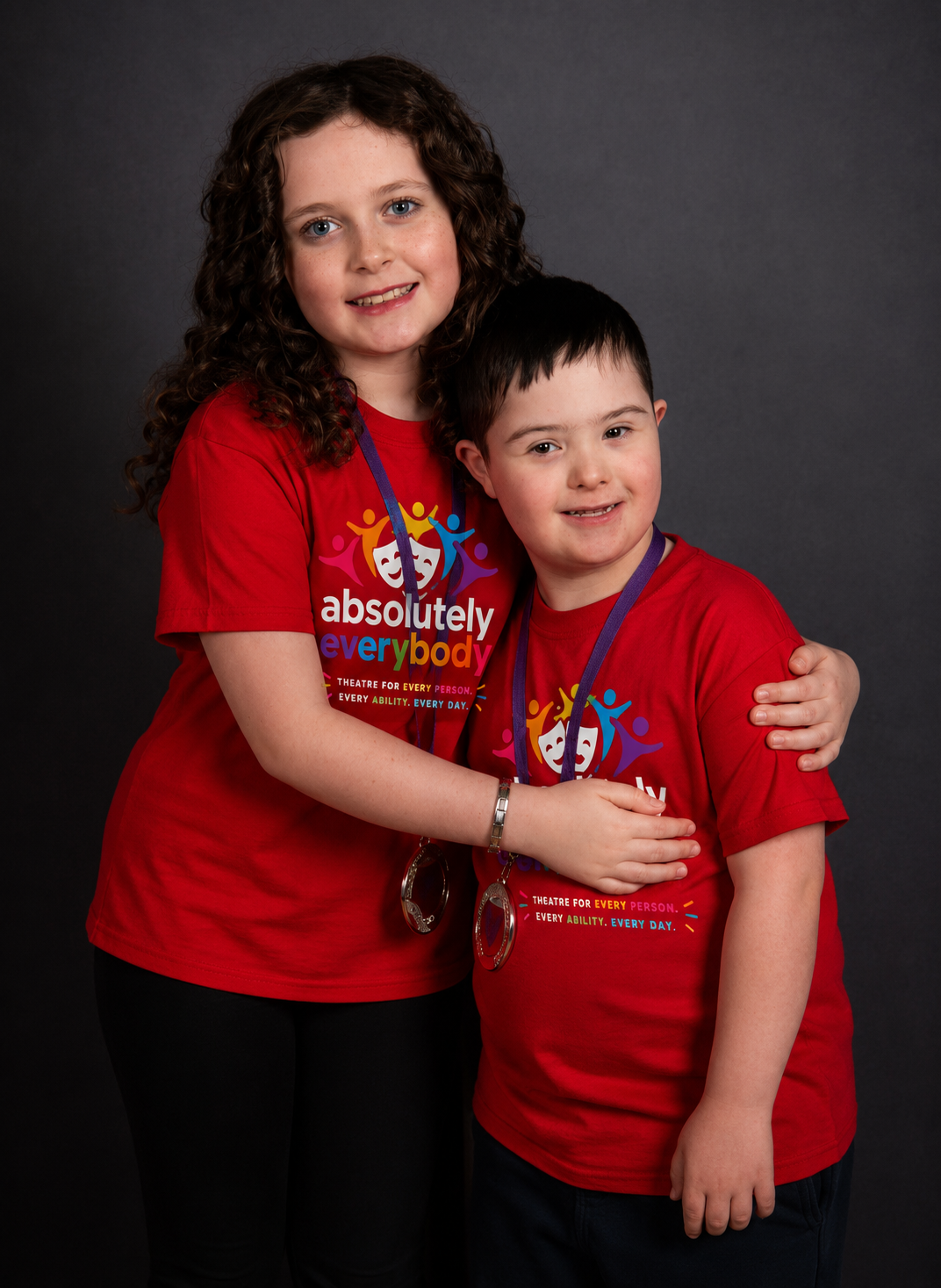 A young girl with curly brown hair and blue eyes hugging a young boy with straight dark hair, both wearing red t-shirts with a design promoting inclusion and ability, standing against a dark background.