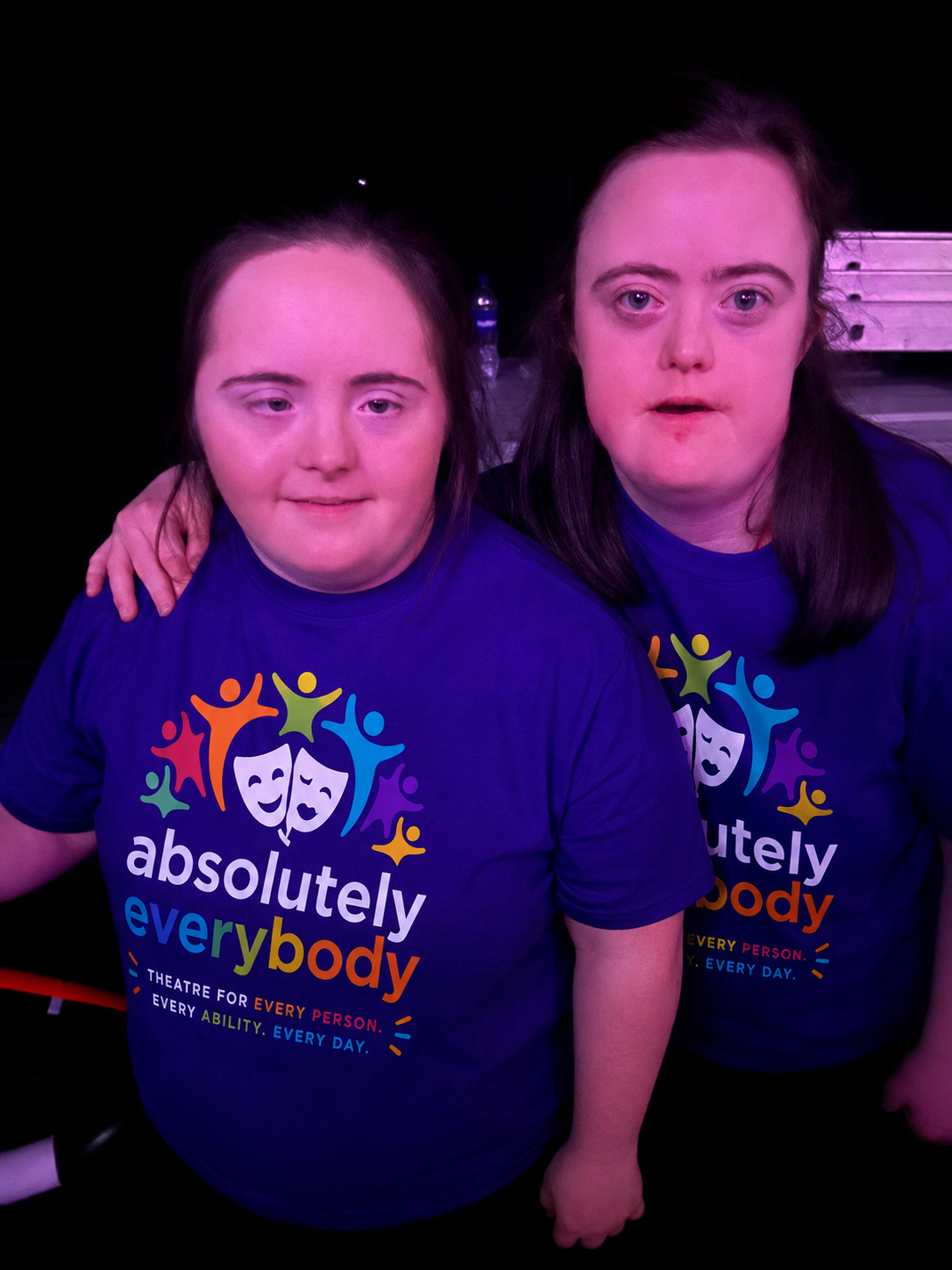Two women wearing matching dark blue t-shirts with colorful logo and text, sitting closely together, one has her arm on the other's shoulder, both have long dark hair, in a dark setting with a black background.