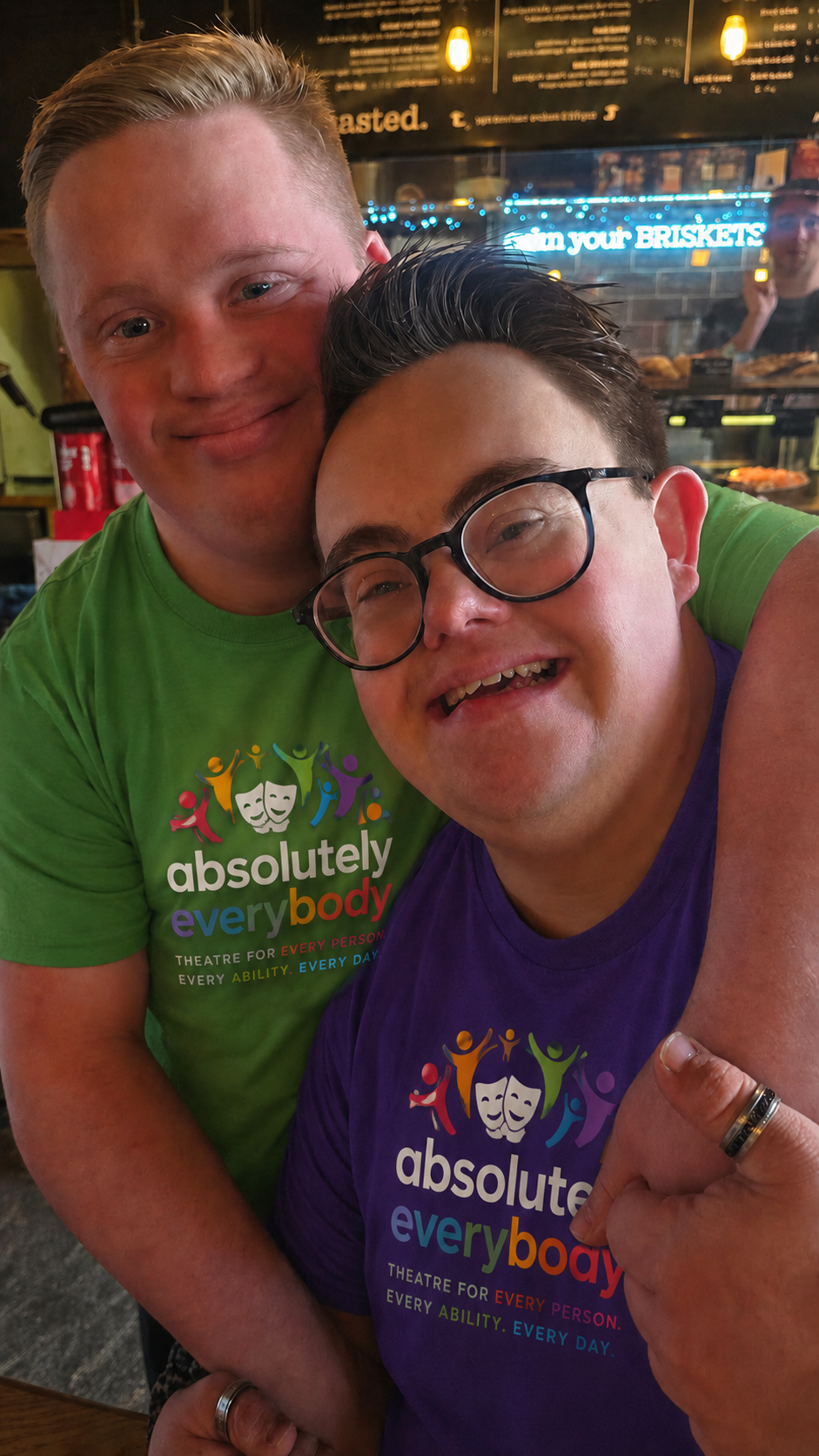 Two smiling men hugging in a restaurant, wearing colorful shirts with the phrase 'absolutely everybody' and graphics of theater masks and diverse people.