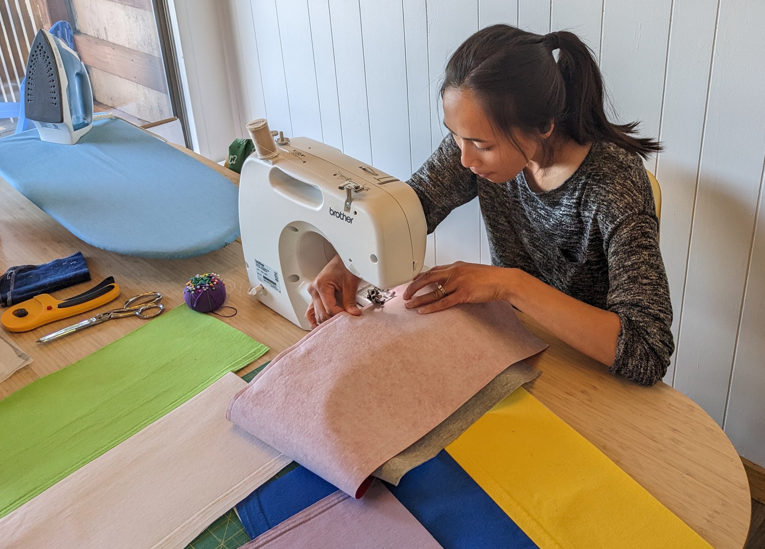 A woman sewing fabric on a sewing machine at a table with sewing supplies and colorful fabric swatches.