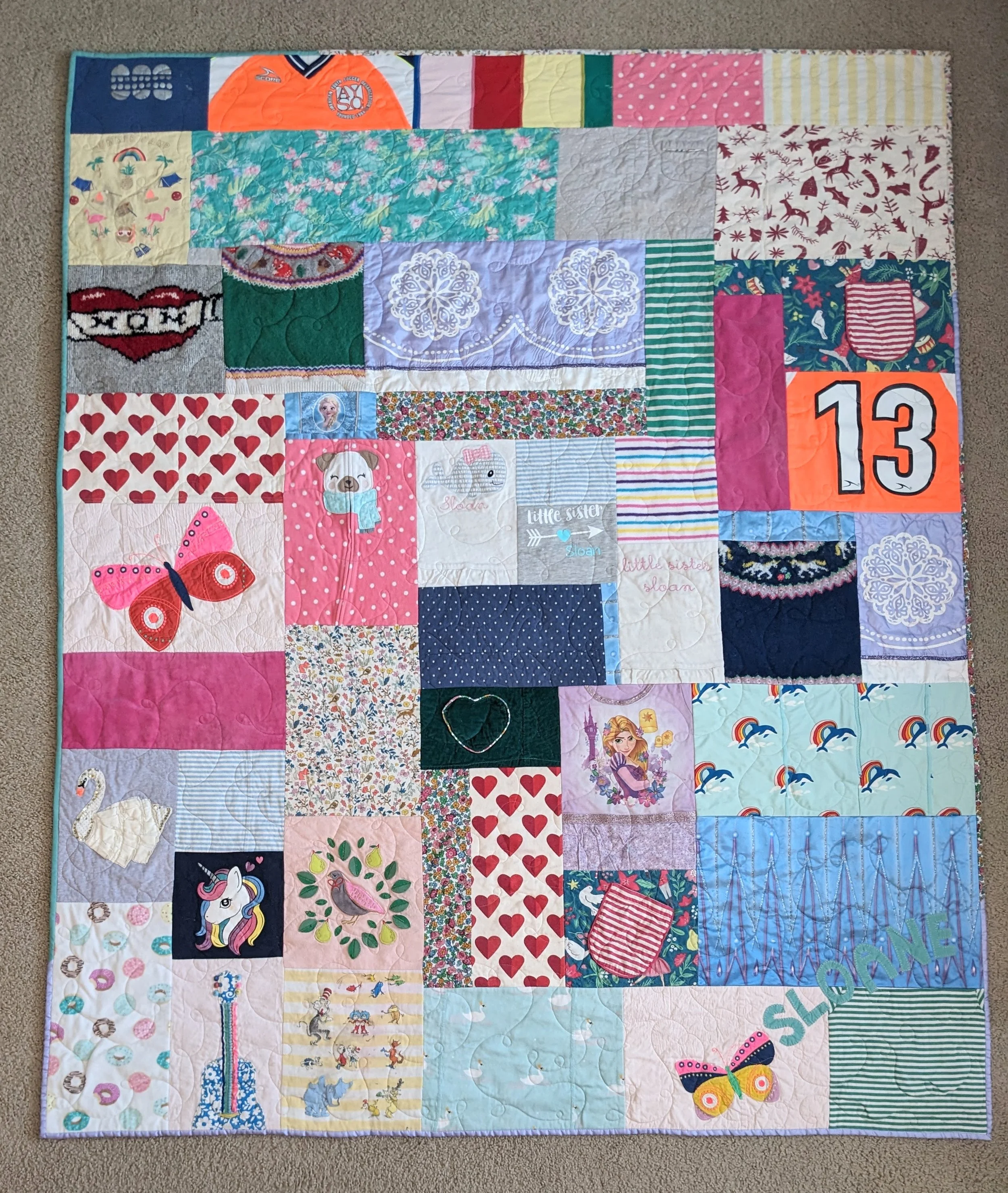 A colorful patchwork quilt with various fabric squares depicting different patterns, characters, and images, including a unicorn, hearts, a butterfly, snowflakes, and a rainbow, laid on a beige carpet.