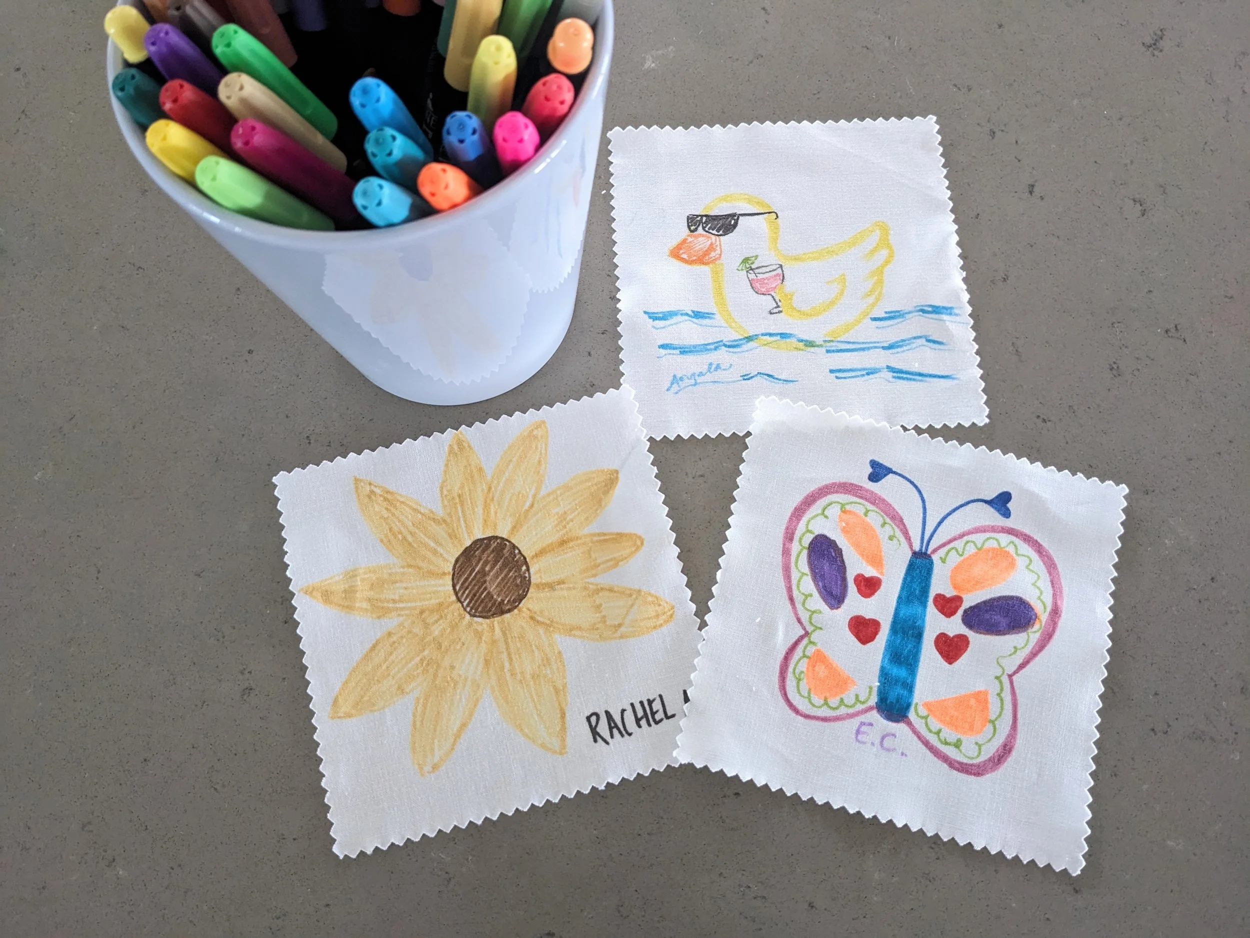 Colorful drawings of a sunflower, a butterfly, and a duck on pieces of fabric, with a cup filled with multicolored markers nearby.