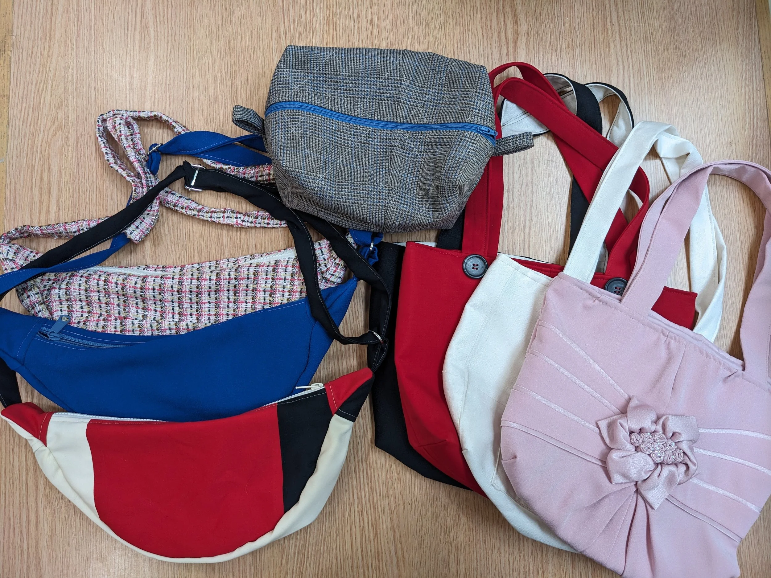 Various colorful handbags and a gray plaid backpack are arranged on a wooden surface.