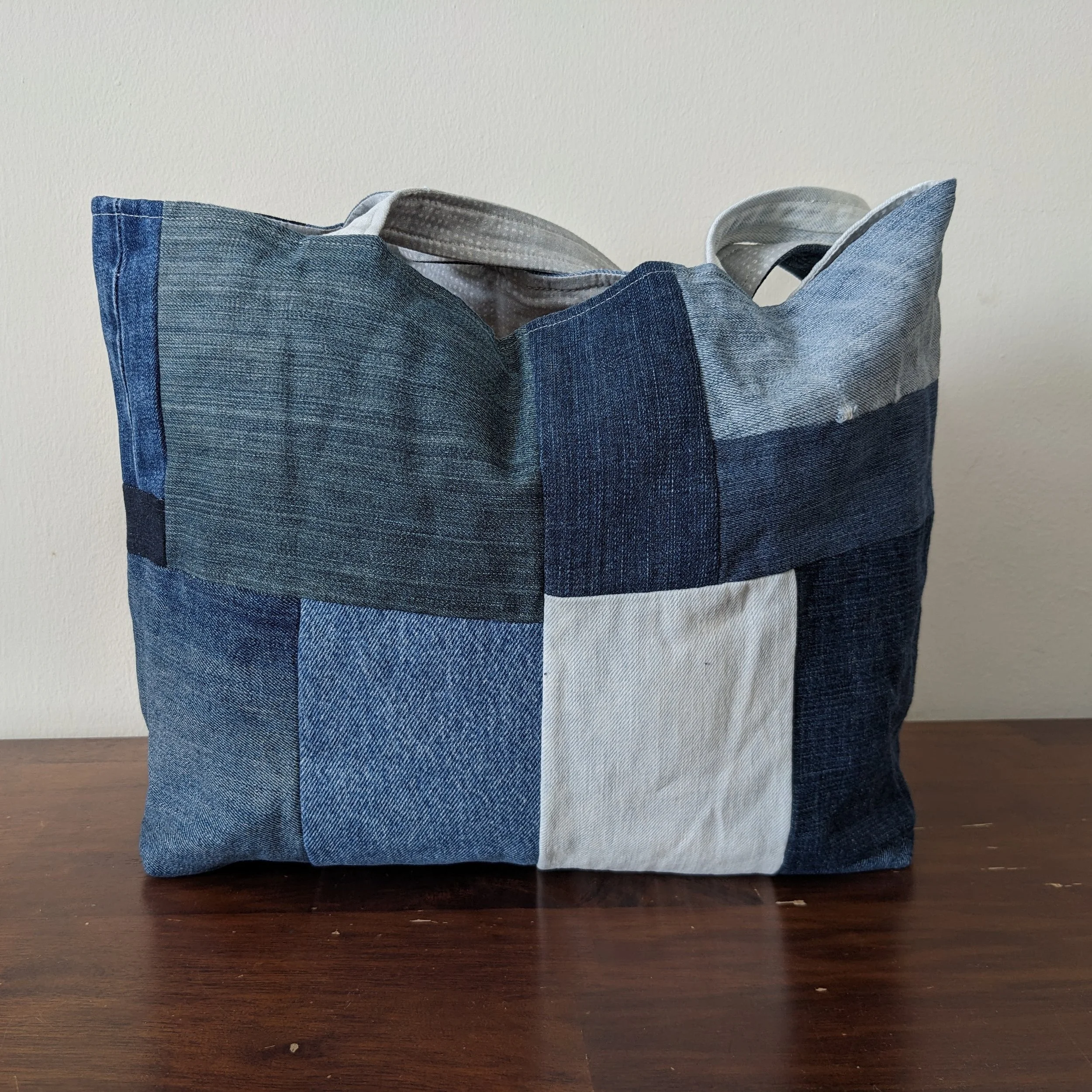 A fabric bag made from blue denim and gray patterned cloth, placed on a wooden surface with a neutral wall background.