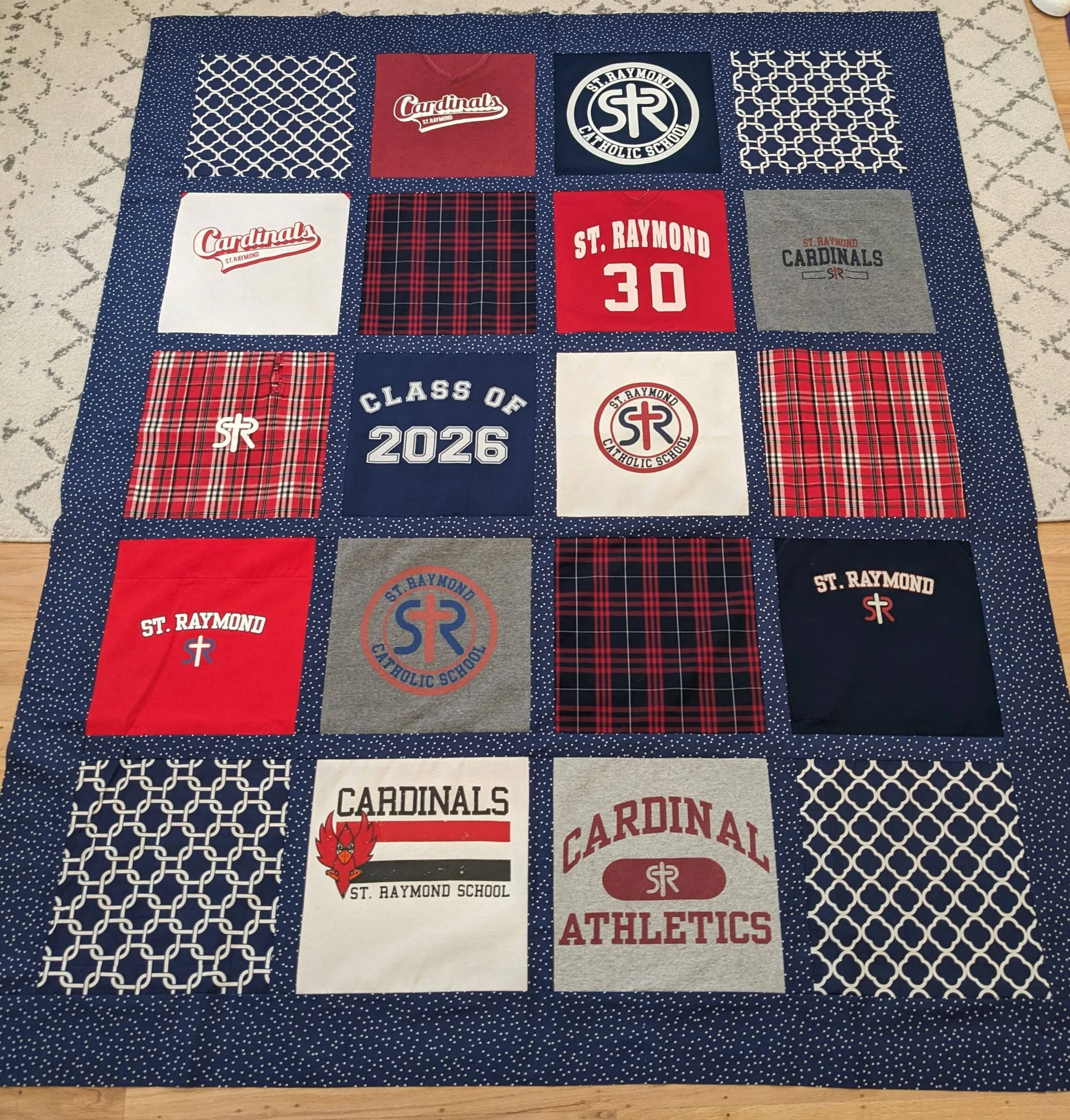 A quilt with a blue background and white polka dots, featuring various school and sports-themed designs, including logos, team names, and slogans for St. Raymond Catholic School, with colors primarily red, white, gray, and navy blue.