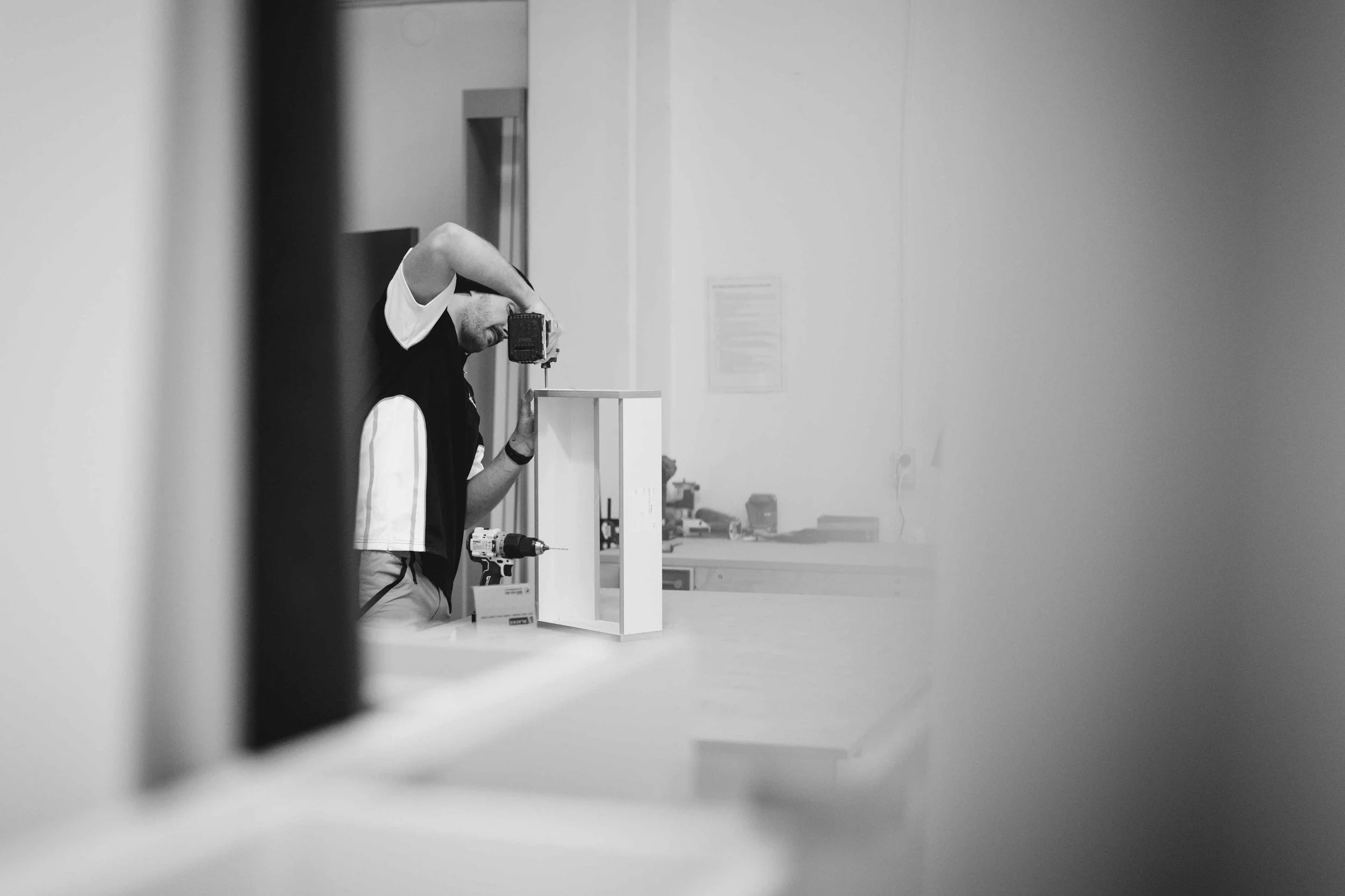 A person working on a small white structure inside a well-lit room, with a drill on the table and a small sign in front, viewed through an opening or frame.