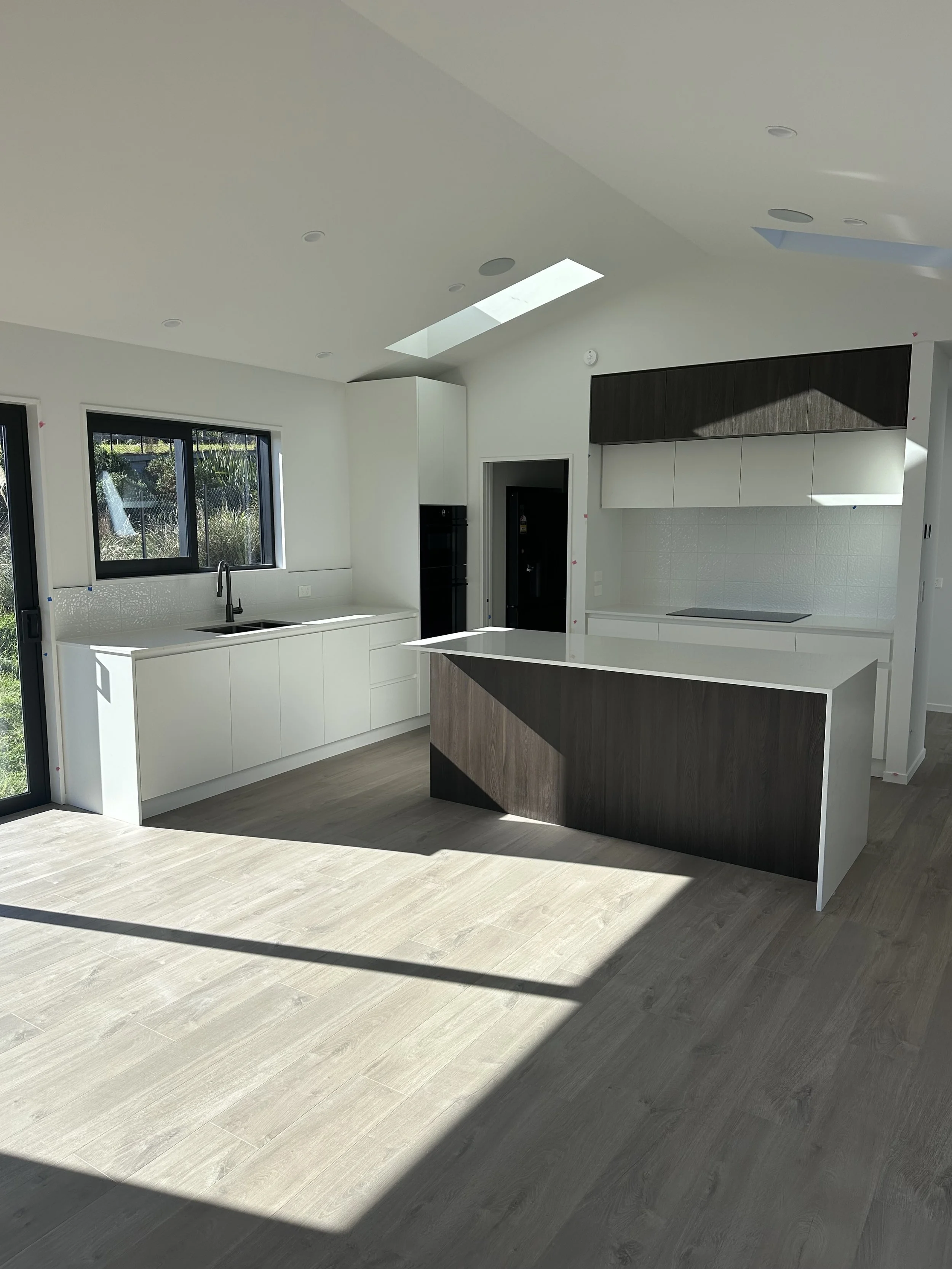 Modern kitchen with white cabinets, black accents, large window, and a skylight, with light wood flooring and sunlight casting shadows.