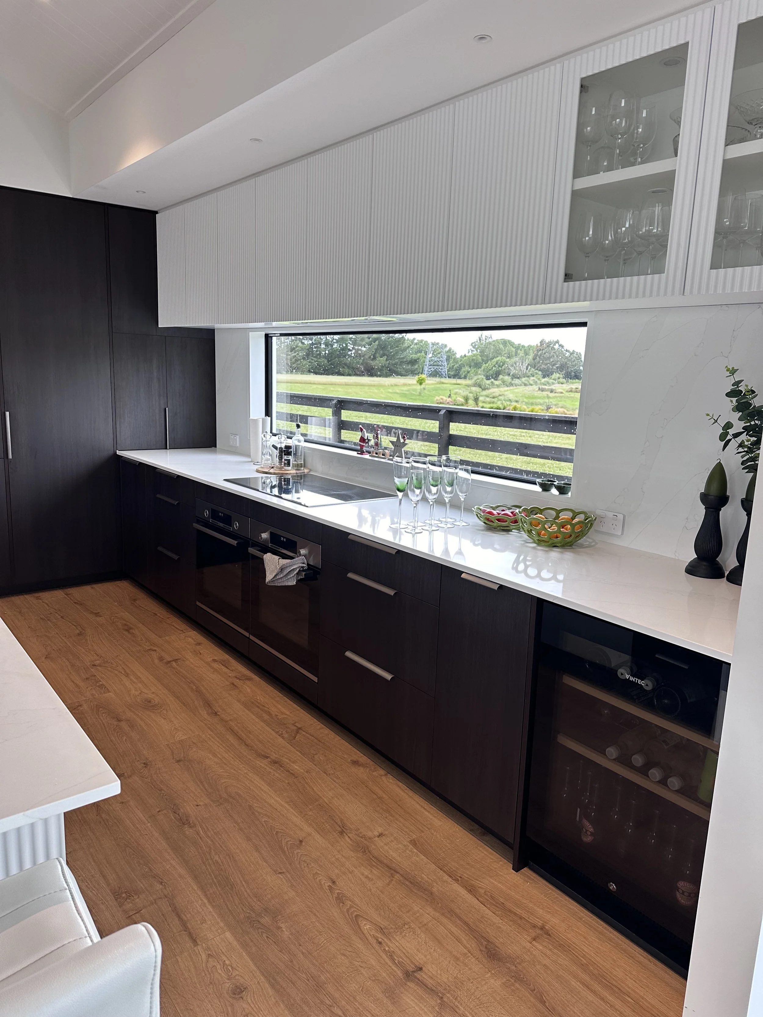 Modern kitchen with dark cabinets, white marble countertop, large window overlooking green landscape, and decorative bowls and glassware on the counter.