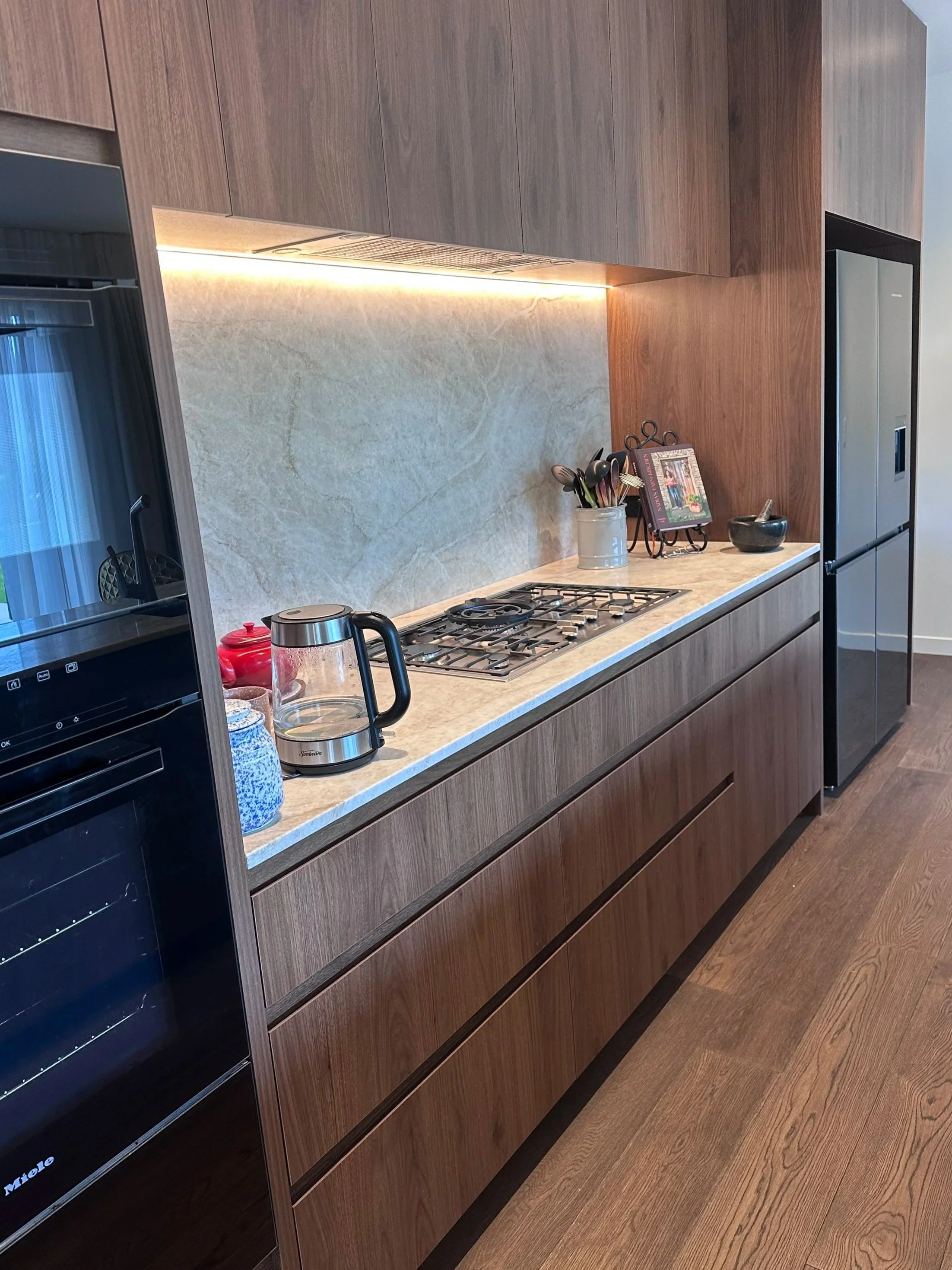 Modern kitchen with wooden cabinetry, beige marble countertop, stainless steel appliances, and a small decorative frame and utensils on the counter.
