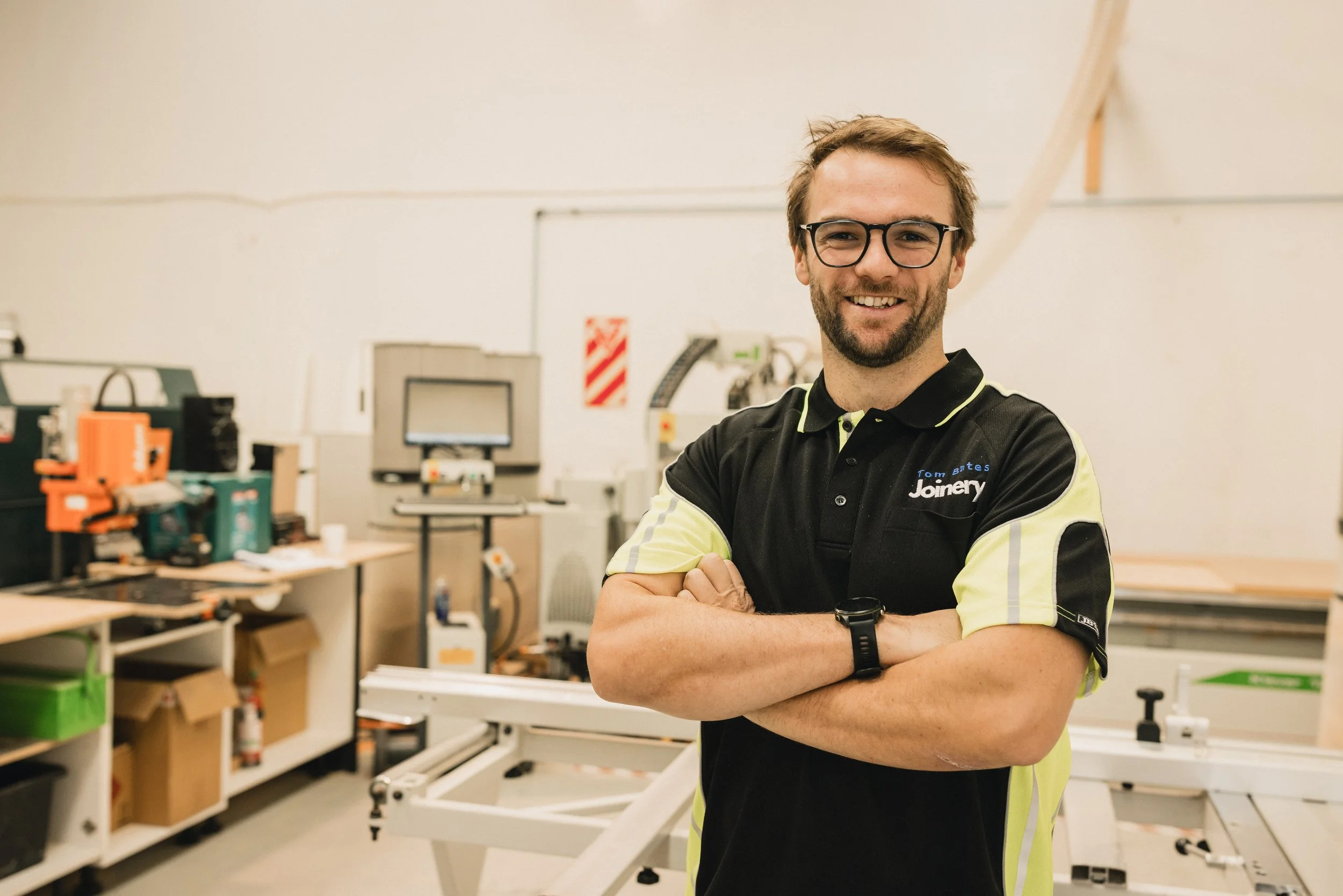 A man wearing glasses and a black and yellow polo shirt with the words "Tom Bates Joinery" smiling with arms crossed in a workshop filled with equipment and tools.