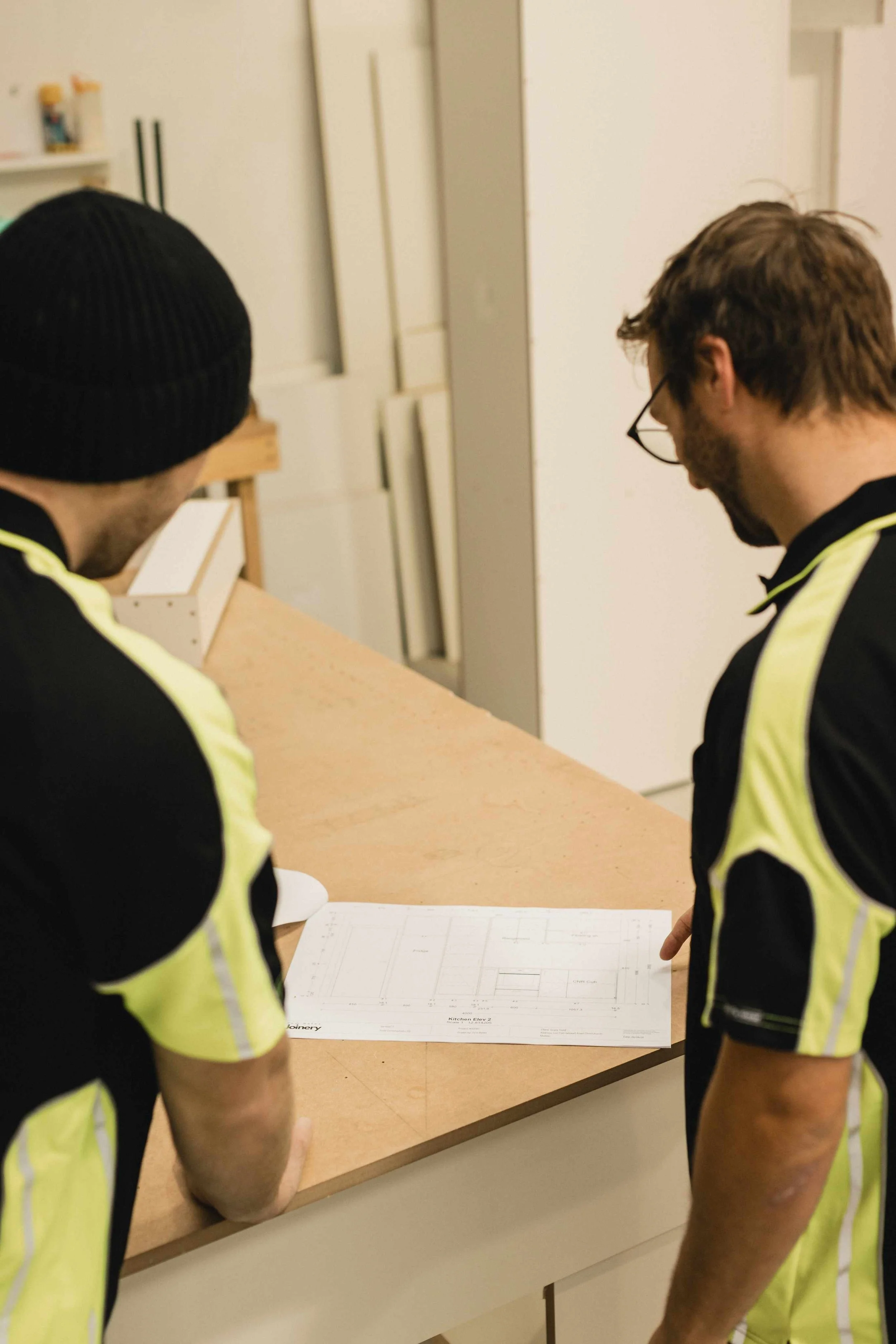 Two men in black and yellow shirts are looking at architectural blueprints on a table in a room with white walls and cabinets.