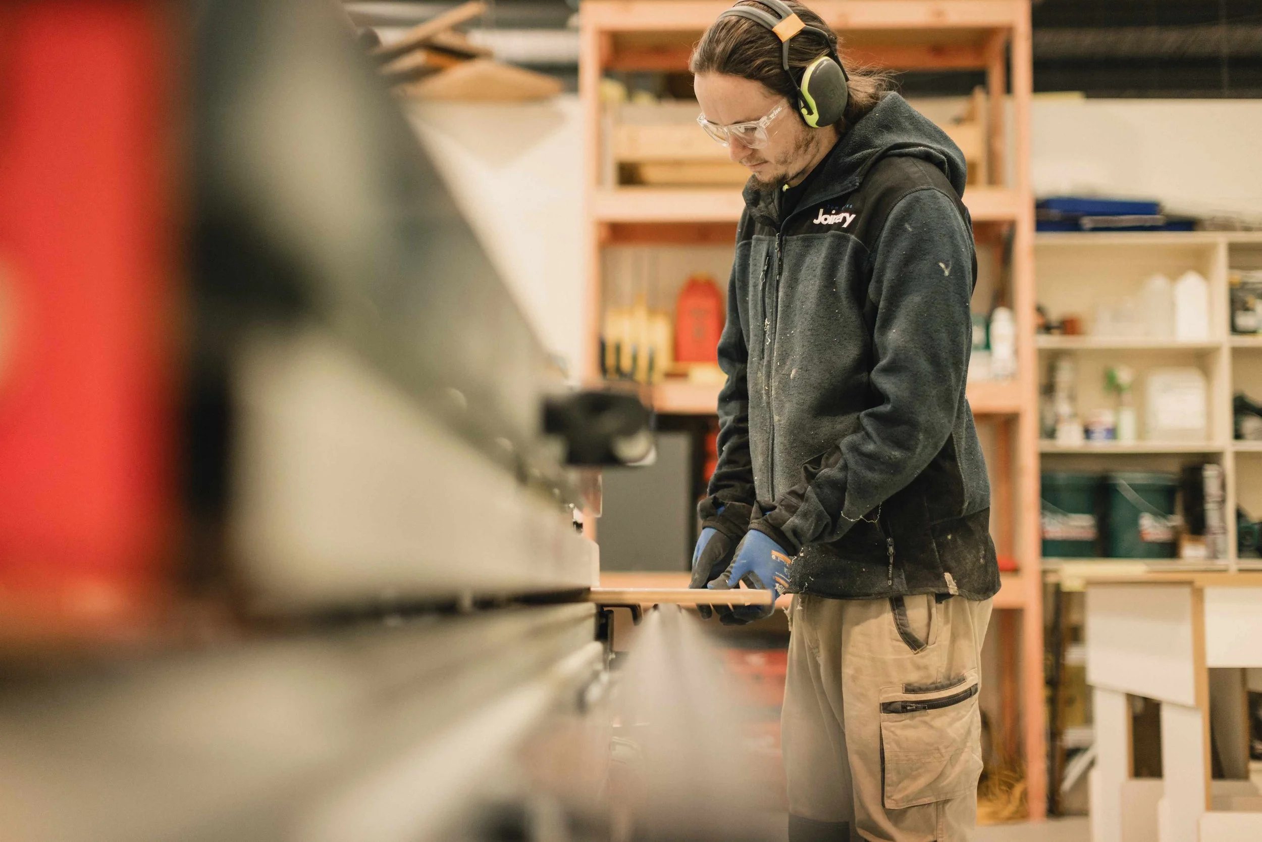 A man wearing safety goggles, headphones, a black jacket with a logo, and work gloves is working in a workshop with wooden shelves and various tools and supplies in the background.
