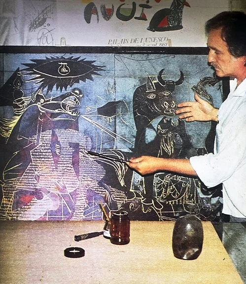 A man in a white shirt pointing to artwork of bulls, a horse, and an eye on a mural in the background.