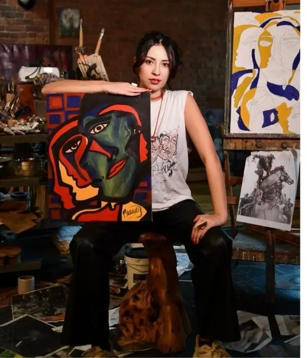 Young woman, artist named Juliana Plexxo, sitting with her right arm resting on a colorful abstract painting of a face, in an artist's studio with various artworks and art supplies around her.