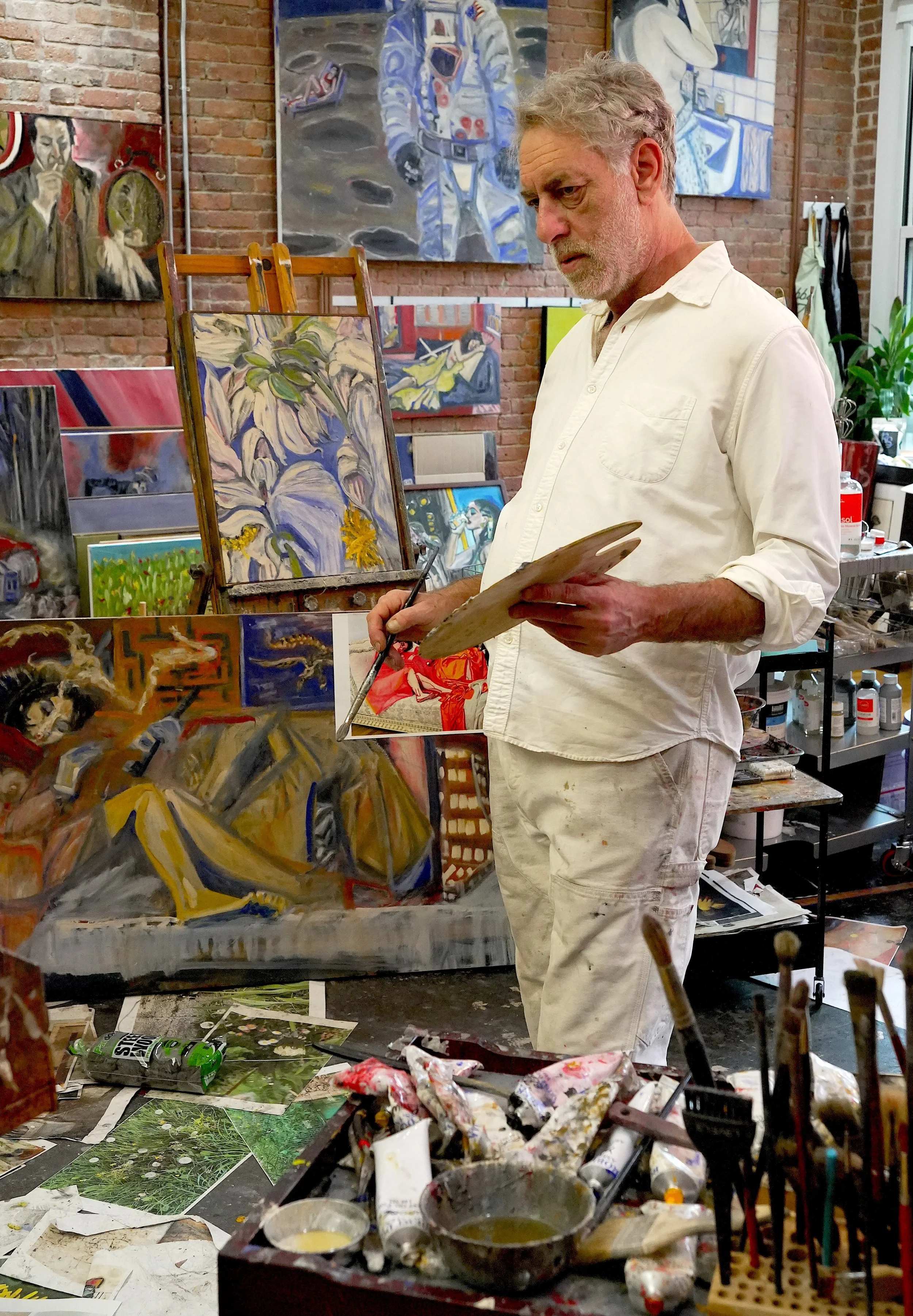An artist stands in a studio, surrounded by paintings of various subjects, wearing a white shirt and paint-splattered white pants, holding a palette and brush, with an art supplies table in front filled with paints, brushes, and paintings on the floor, brick walls, and numerous artwork displayed behind him.