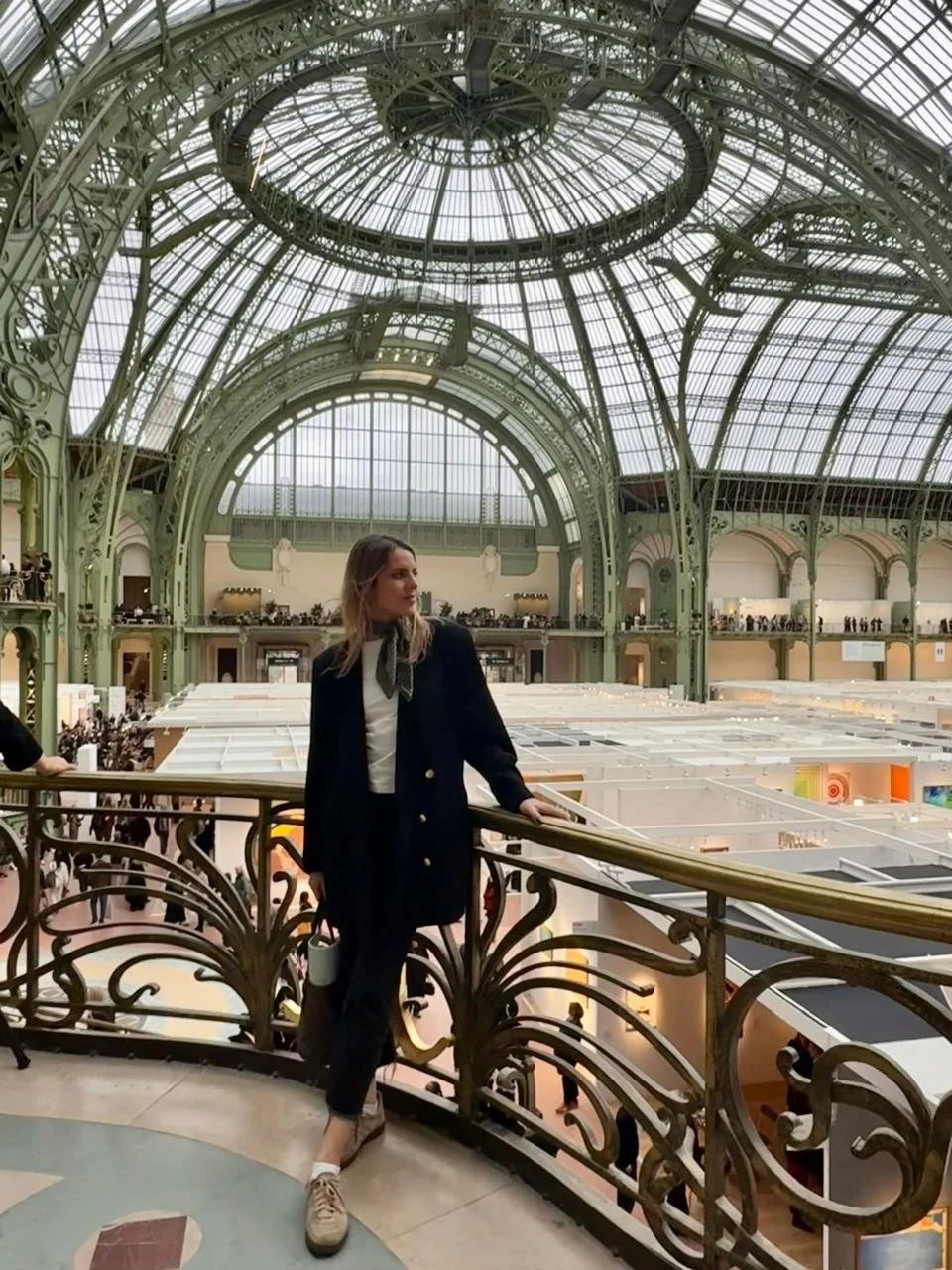 A woman in formal attire stands on an ornate balcony inside a large glass and metal-domed building, overlooking an expansive interior space filled with galleries and crowds.