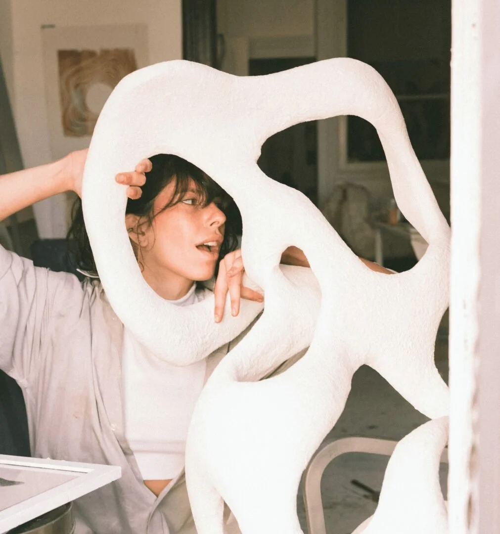 A woman with dark hair and bangs looking through a large, abstract, white sculpture with holes. She is indoors, wearing a light-colored, long-sleeved top, and appears to be examining or posing with the sculpture.