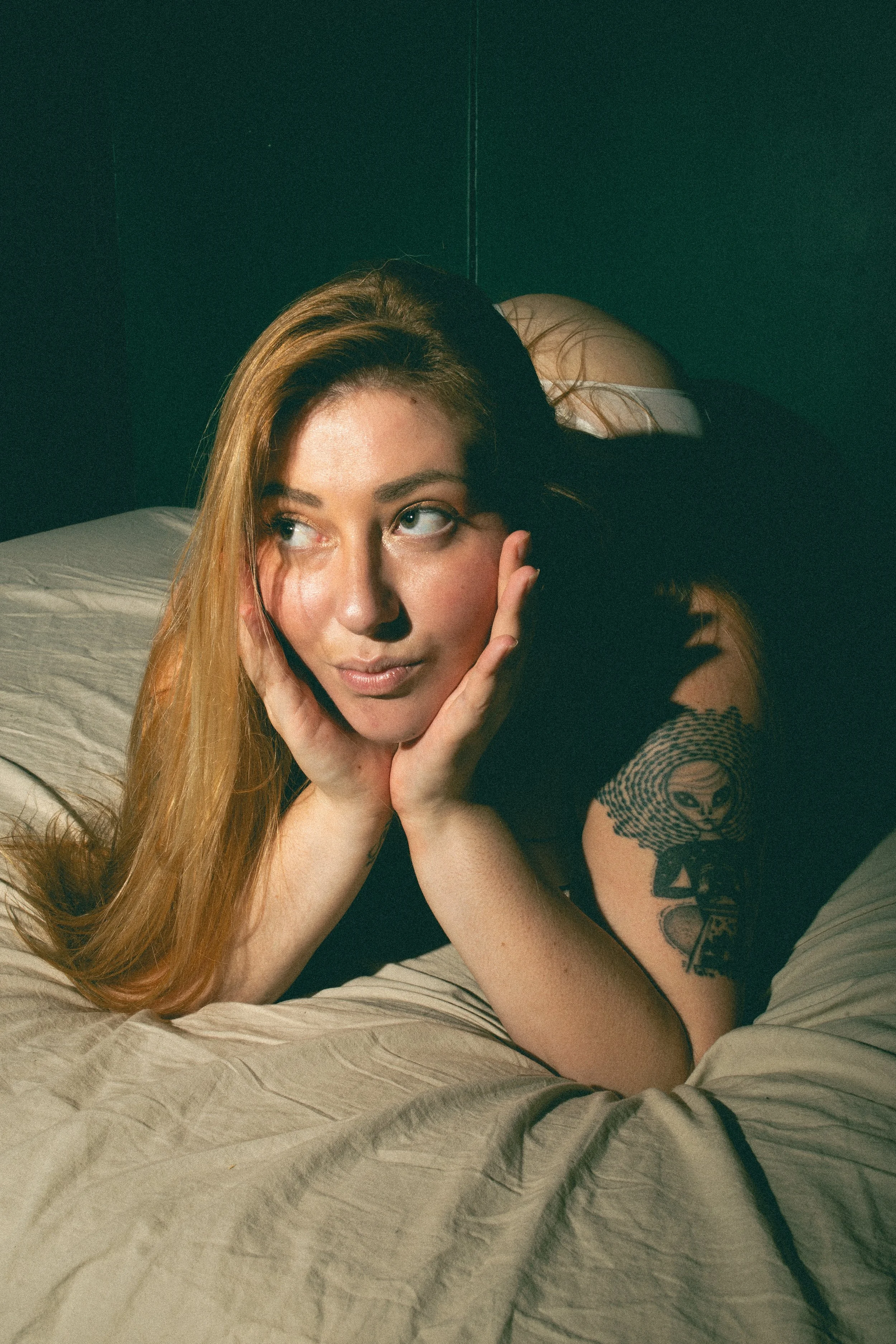 Jeune femme aux cheveux longs et blond, posant sur un lit avec un fond noir, portant un t-shirt noir, regardant par-dessus son épaule, avec tatouages sur son bras.